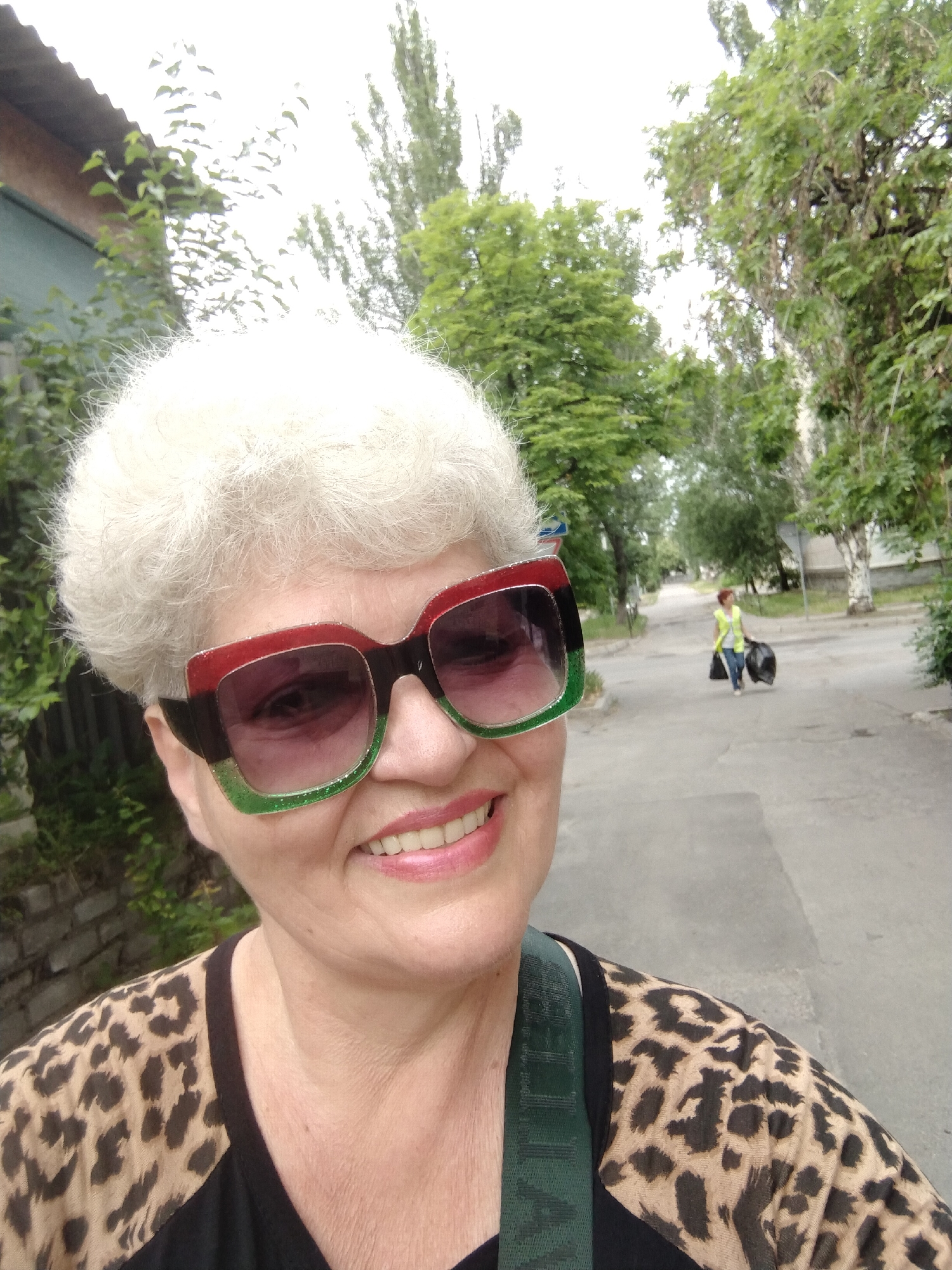 A woman with short, white hair and oversized sunglasses with red and green frames smiles for a selfie outdoors. She is wearing a leopard-print top. In the background, a street is visible with trees lining the sides. A woman in a yellow vest is walking away in the distance carrying two black trash bags. The scene appears to be in Horishni Plavni, Ukraine.