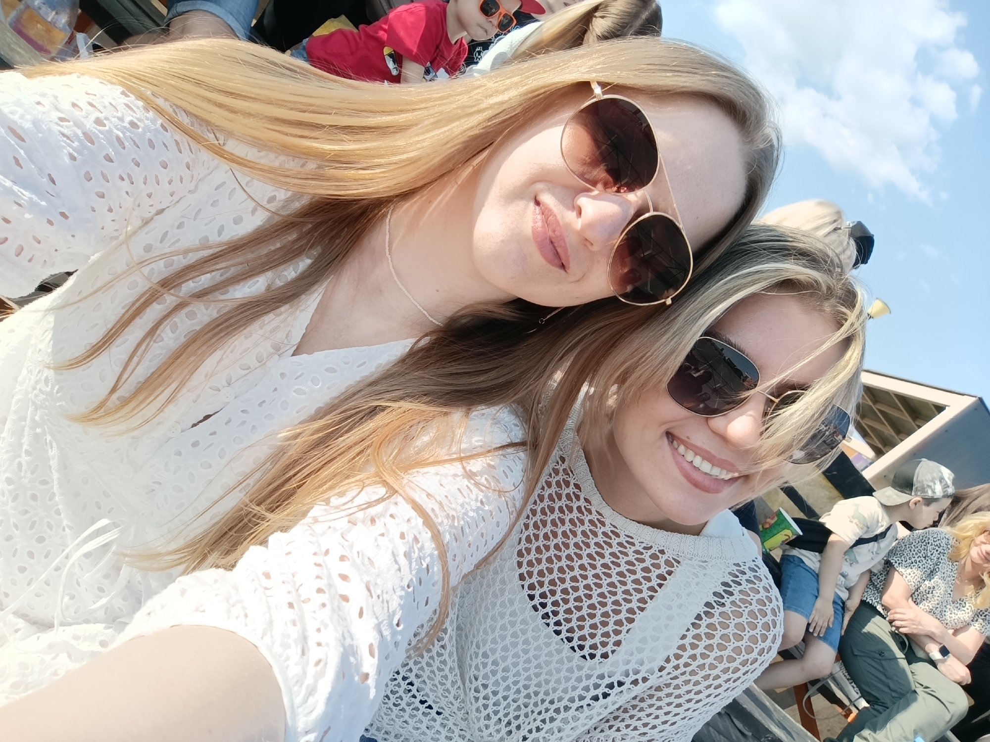 Two women are posing close together, presumably for a selfie. The woman on top has long, blonde hair and is wearing round, aviator-style sunglasses with a gold frame. She is wearing a white, eyelet-style dress. The woman below has blonde, highlighted hair and is wearing dark sunglasses. She is wearing a white, net-style shirt over a black top. There are other people in the background, including a child in a red shirt and sunglasses. The sky is visible in the upper right corner. The location appears to be outdoors in an unspecified city.
