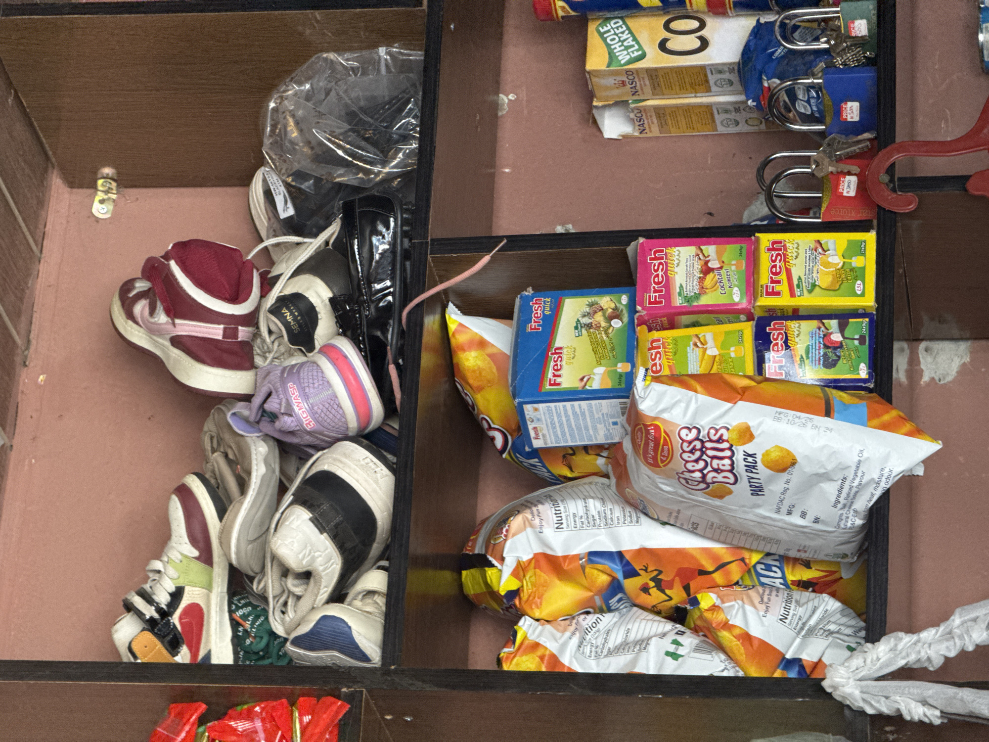 This image shows a close-up, top-down view of a shelf in a store, likely in Abuja, Nigeria, filled with various items. The shelf is divided into compartments, with the left side primarily containing shoes and a plastic bag. On the right side, there are rows of boxed juices and bags of chips, along with padlocks and keys.
The shoes appear to be a mix of sneakers and possibly some casual footwear, in various colors and styles. Among them is a distinctive pair of high-top sneakers with maroon and white accents. There's also a clear plastic bag containing what looks like more shoes or accessories.
The right side of the shelf is more colorful, with bright packaging for juices and snacks. Boxes labeled "Fresh quick" in flavors like "Cocktail Kokteyl," "Banana," and "Lemon Citron" are stacked neatly. Bags of "Cheese Balls PARTY PACK" are also prominent. Interspersed with the products are several padlocks, two blue and one red, some with keys attached. The red padlock has a label indicating "PRICE N30".
In the background, there are boxes of "NASCO WHOLE FLAKED Co" cereal. The overall impression is of a well-stocked, perhaps slightly cluttered, retail space. The lighting suggests an indoor setting, likely during daylight hours, given the absence of artificial lighting glare. There are no people visible in the image. The text elements include product names, nutritional information, pricing, and manufacturing details.