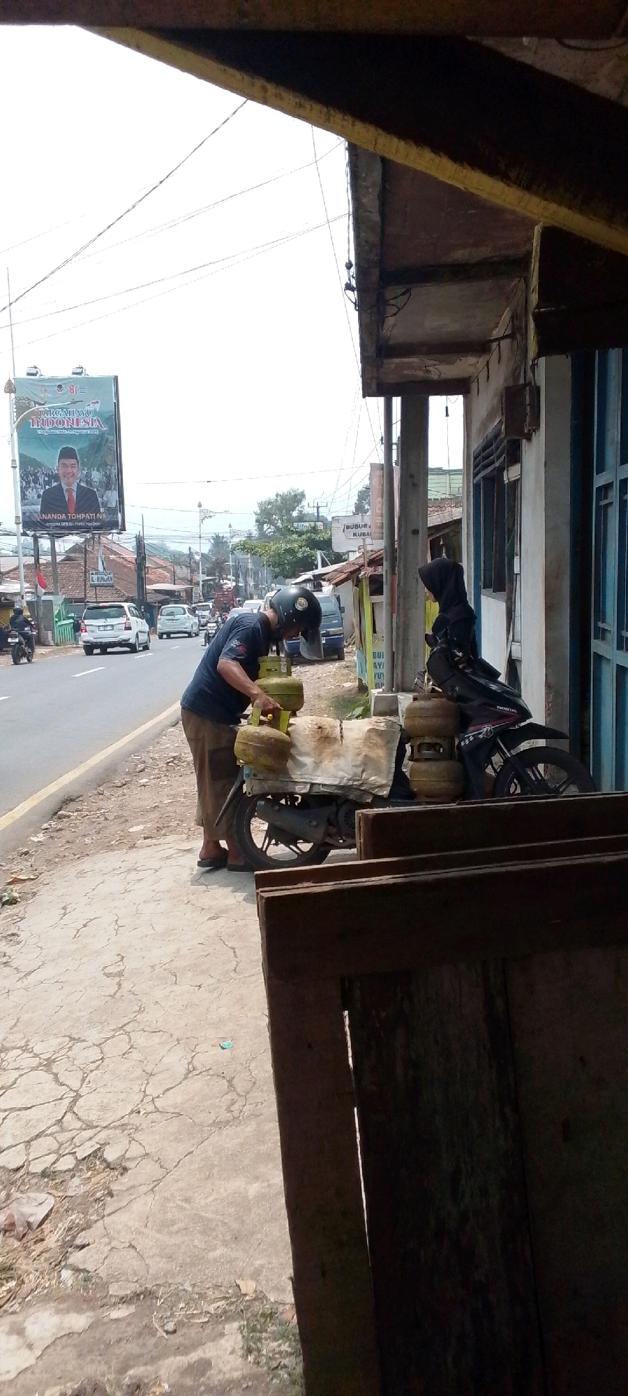 In the city of Cilaku, Indonesia, a man wearing a dark blue shirt, beige pants, and a black helmet is seen from the side, leaning over a motorcycle. The motorcycle is carrying several green gas cylinders, some of which are wrapped in a light-colored tarp. The man appears to be handling one of the gas cylinders. To the left of the motorcycle, a busy street is visible with cars and motorcycles driving on it. A billboard with an image of a man in a suit and text in Indonesian is positioned on the left side of the street. The scene is bordered by a building on the right with blue window frames and wooden structures in the foreground. The ground consists of cracked concrete and dirt.
