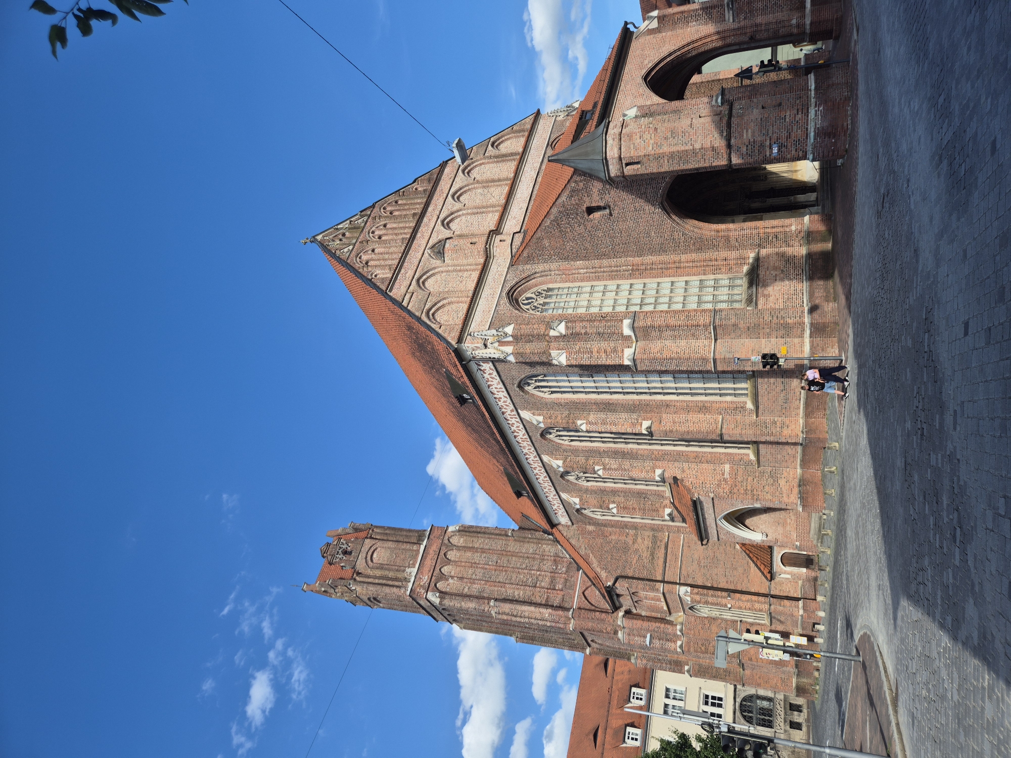 The image shows the St. Martin's Church in Landshut, Germany, a Gothic brick church with a tall, slender tower and a red tiled roof. The church is made of red brick and features arched windows and entrances. Two people are walking on the cobblestone street in front of the church. The sky is blue with scattered white clouds. The scene appears to be a sunny day.