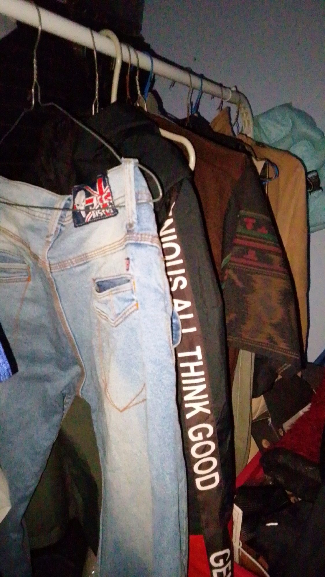 The image shows a close-up view of clothing items hanging on a rack. The foreground features light-wash denim jeans with a patch on the waistband. To the right of the jeans, a black garment with white text vertically printed on it reads "VICIOUS ALL THIN K GOOD GE". Further to the right, other clothing items in brown and tan hues are visible, as well as a light blue garment. The items are hung on a white rod supported by hangers. The background is dark and out of focus, with what appears to be a wall or similar surface. The image does not provide any specific information or context that would allow for identification of a location in Cintaratu, Indonesia.