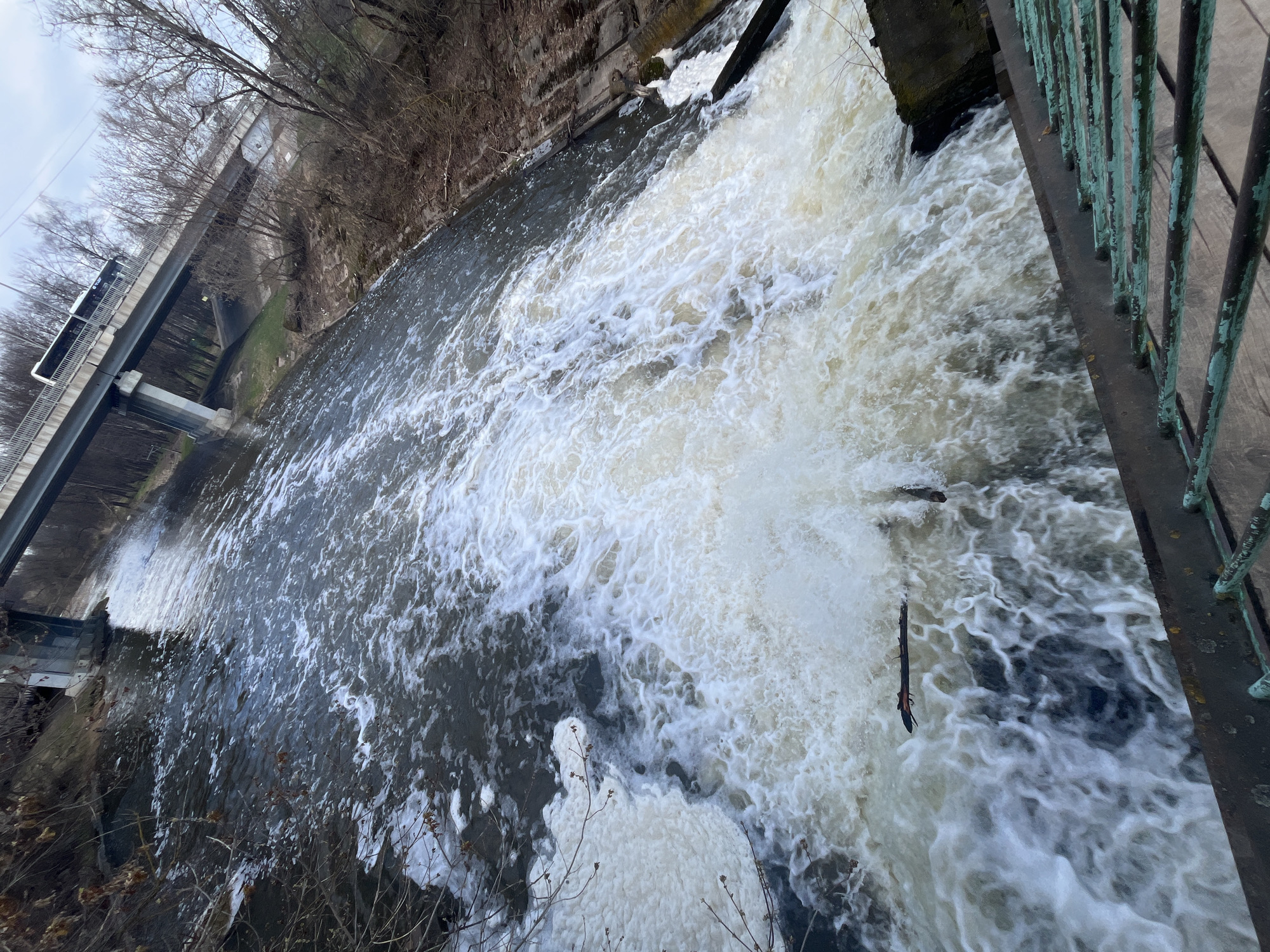 The photograph, taken from an elevated vantage point, presents a view of a waterfall and a section of the Sestra River in Klin, Russia. To the left, a bridge can be seen, with a light-colored bus or van positioned on it. The river flows from left to right, culminating in a cascade over a weir or dam. The water is turbulent and frothy where it descends. A weathered metal railing appears on the right edge of the frame. The banks are lined with trees without leaves. The sky is overcast.