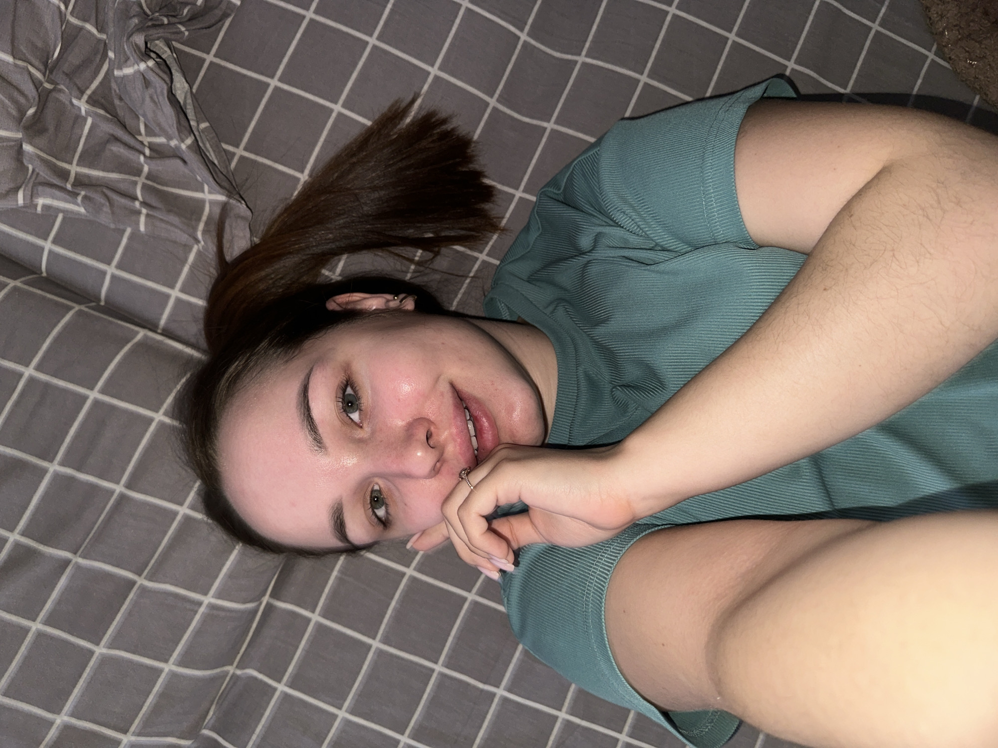 A young woman with fair skin and brown hair tied back in a ponytail lies on a gray and white checkered surface, possibly a bed or blanket. She is wearing a short-sleeved teal shirt. Her right hand, adorned with a ring, is near her mouth. The background is nondescript, with a glimpse of a textured surface in the upper right corner and rumpled fabric on the left. No information suggests the location is Chaikovsky, Russia.