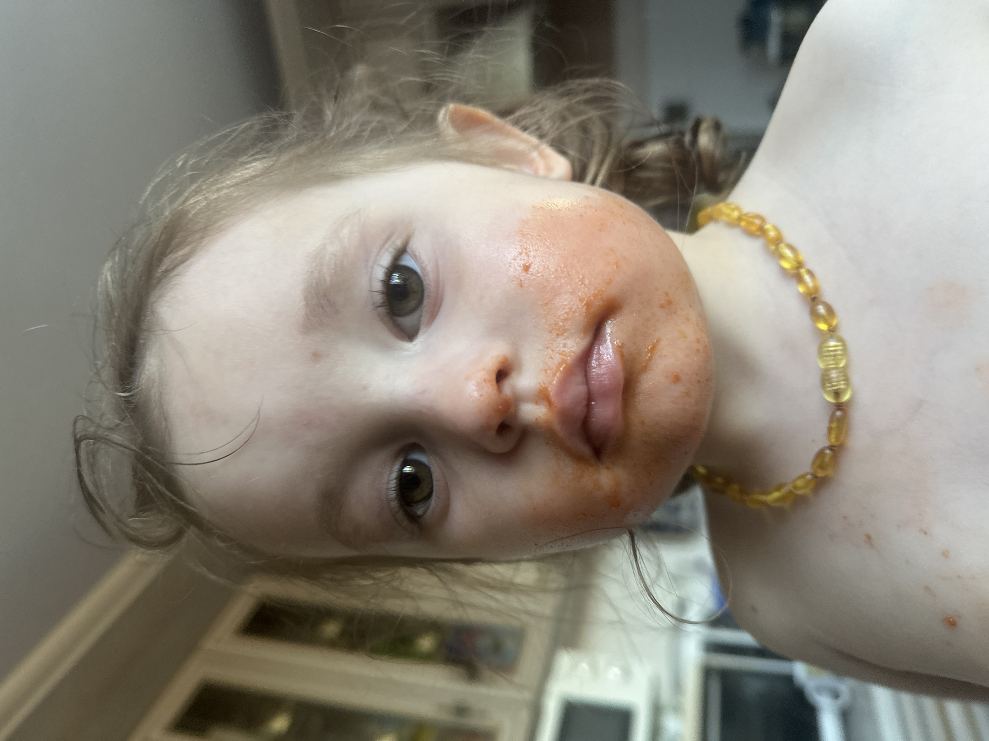 The media file is a close-up, slightly tilted photograph of a young child's face and upper body. The child has light brown hair, which is slightly messy, and large, dark eyes. Their face is smeared with a reddish-orange substance, likely food, around their mouth and on their cheeks. The child is wearing a necklace made of translucent, amber-colored beads. The background is blurred, but appears to be an indoor setting with what looks like cabinets or furniture visible. The lighting suggests it could be daytime. There is no visible text in the image. The overall impression is of a candid moment, perhaps after a meal.