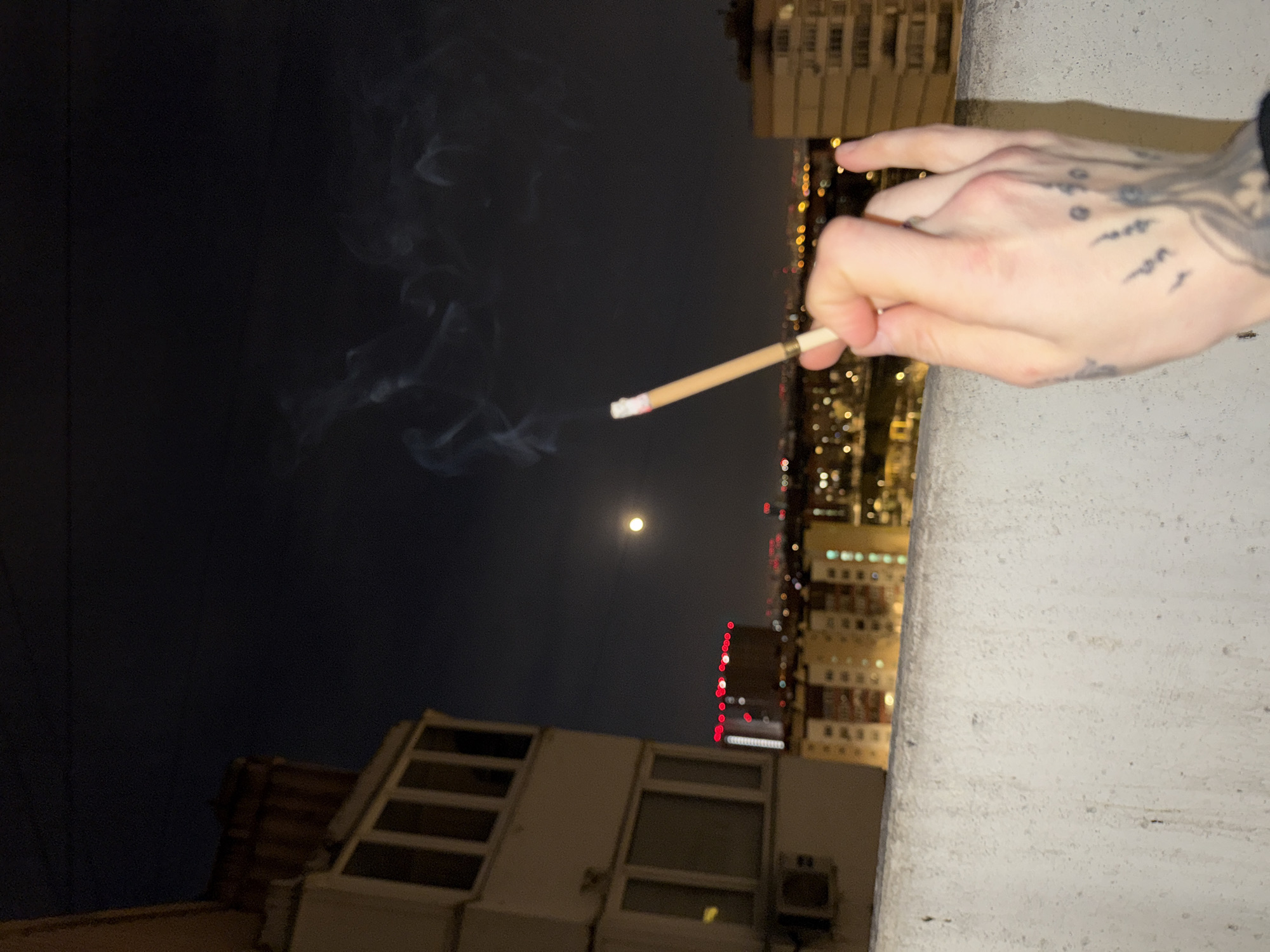 A person with tattoos on their hand holds a lit cigarette, smoke rising from it. Behind them is a nighttime cityscape with buildings and lights. The moon is visible in the sky. The scene is in Moscow, Russia, indicated by the style of architecture and the presence of buildings with visible Cyrillic characters on some of the buildings.