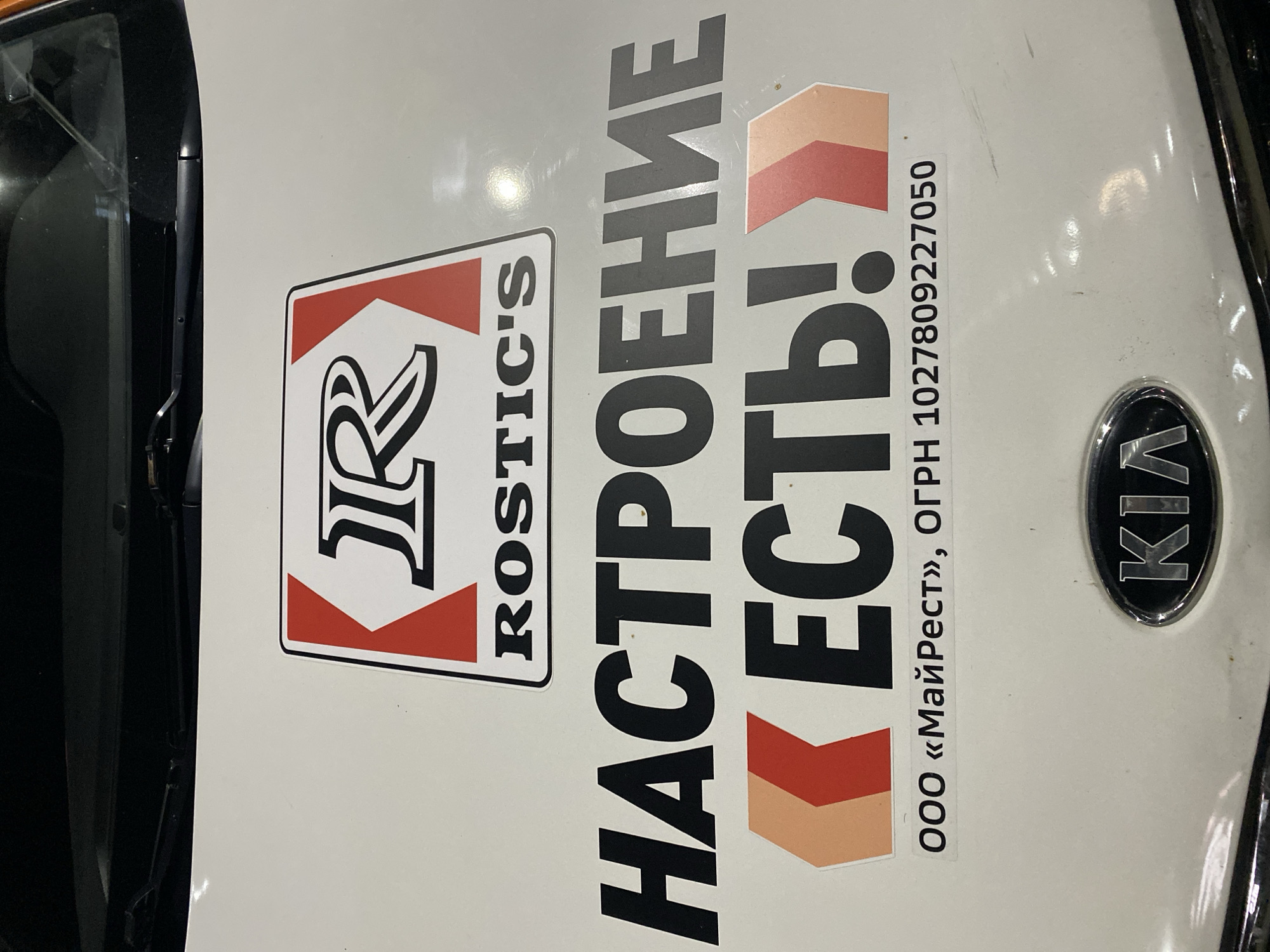 This image displays the white hood of a vehicle, angled and rotated clockwise. A black KIA emblem is visible on the right side of the hood. The hood features several decals, primarily text and logos.
On the left, a distinctive logo features stylized letters, possibly "R R", enclosed within a white rectangle with red chevron-like elements at the top and bottom, reminiscent of the Marlboro cigarette packaging design. Vertically aligned next to this is the word "ROSTIC'S" in black uppercase letters.
To the right of this logo, large black Cyrillic text reads "НАСТРОЕНИЕ ЕСТЬ!", which translates to "Mood is good!" or "There is mood!". Below this slogan, smaller text states "ООО «МайРест»" (LLC "MyRest") and "ОГРН 1027809227050", which is a Russian company registration number.
The vehicle's hood has a glossy white finish, reflecting ambient light, which suggests an indoor setting with artificial lighting or an outdoor nighttime scene. Faint scratches are visible on the white surface. No people or activities are present; the image is a static shot of the vehicle's branding. The branding indicates the vehicle belongs to a company associated with "Rostic's" and "MyRest", likely a commercial or delivery vehicle.