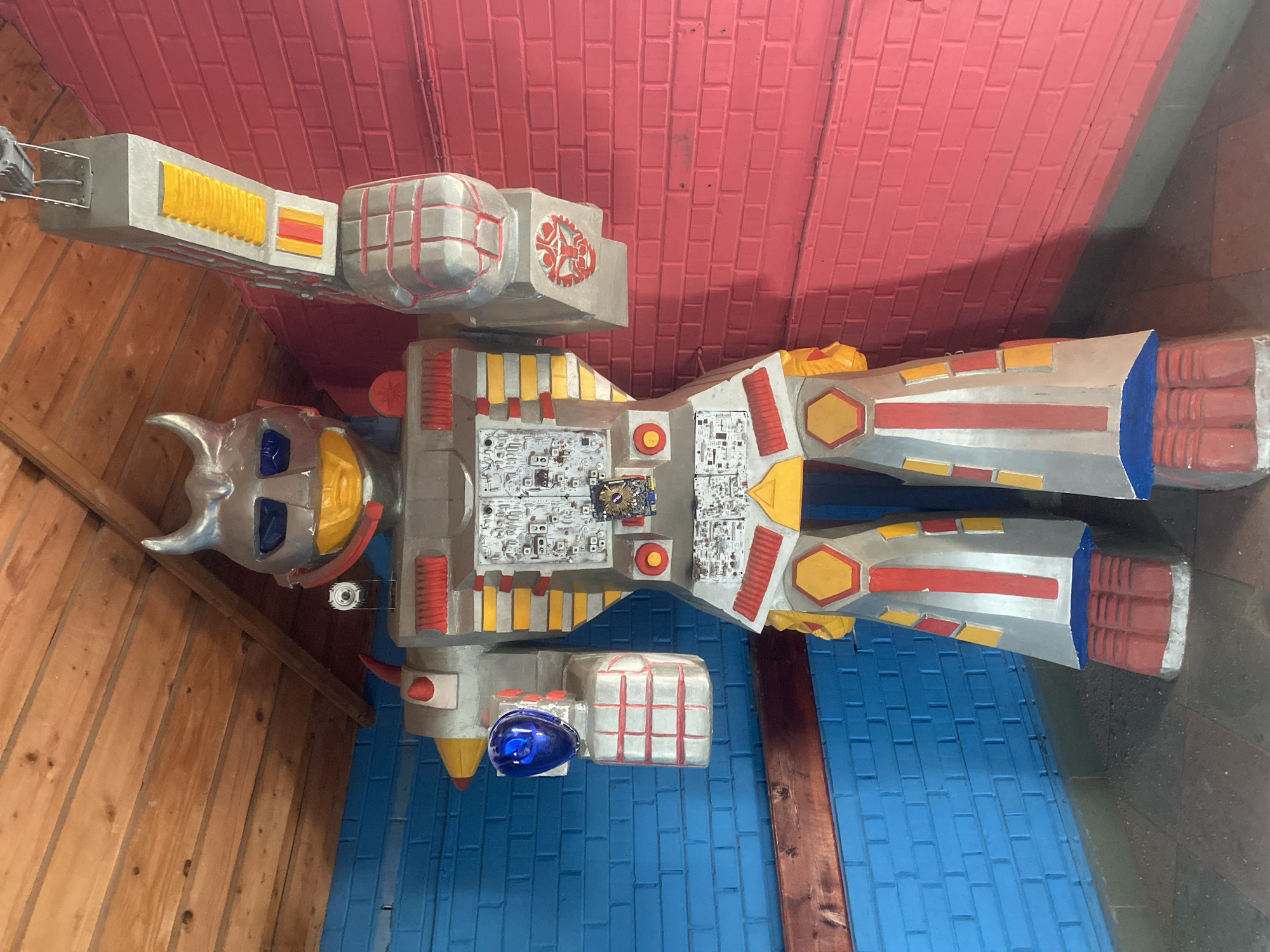 The image displays a large, elaborate robot-like figure, presented as a sculpture or prop in an indoor setting. The robot is primarily silver with striking accents of red, yellow, and blue.
The figure's head is silver, featuring horns, blue eye slots, and a yellow faceplate. Its torso is complex, showcasing what appear to be exposed circuit boards, replete with various small components, indicating a detailed mechanical aesthetic. One circuit board has a small blue component that might be an Intel CPU chip, though any text or logo on it is indistinct. The arms and legs are segmented, adorned with bold red and yellow striped patterns. One arm includes a prominent, dome-shaped blue light or feature.
The background consists of brightly painted walls and wooden paneling. The upper portion of the background is a solid red painted brick wall, while the lower section is a blue painted brick wall. To the left of the figure, there is natural wooden paneling, possibly a ceiling or an angled wall.
The robot appears to be a stationary display, possibly mounted or hanging, with no active event taking place. The overall design evokes a vintage "super robot" style, reminiscent of classic sci-fi or anime characters. The intricate details, especially the simulated electronic components, enhance its mechanical and technological theme. The scene is brightly lit, emphasizing the vivid colors and textures of both the figure and its surroundings. No clearly readable text is visible on the figure or in the background.
