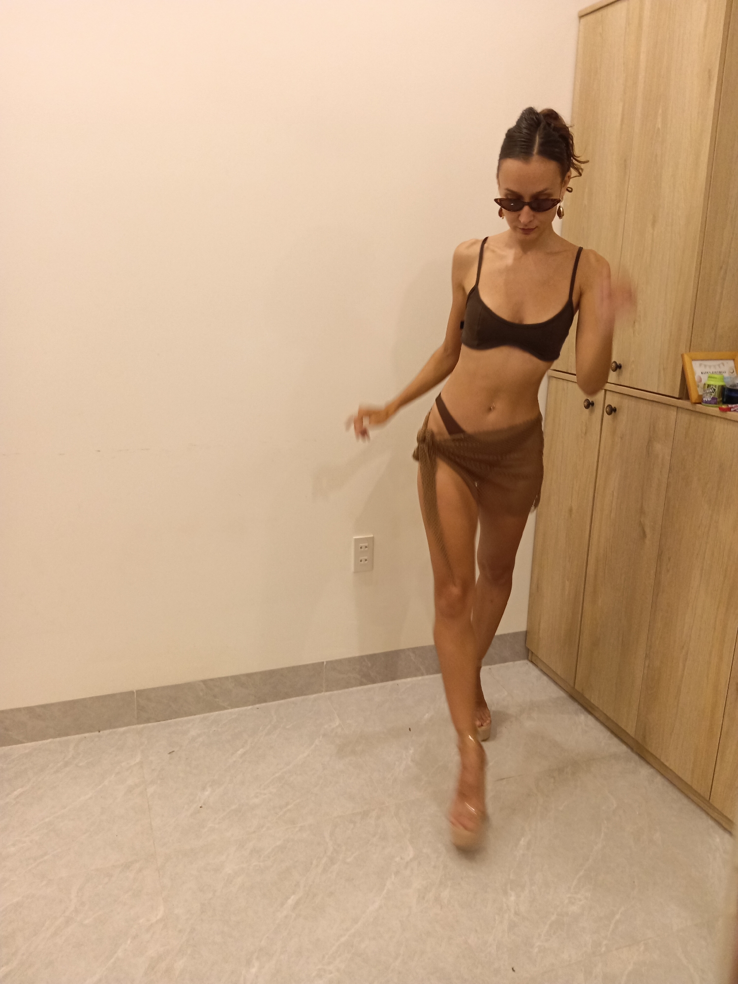 The image features a young woman captured in an indoor setting, likely in Mũi Né, Vietnam. She is actively walking or posing, her body angled towards the right side of the frame, with a slight blur on her right arm suggesting movement.
The woman is wearing a dark brown string bikini top and matching high-cut bikini bottoms, layered with a sheer, textured brown sarong tied around her waist. Her accessories include dark, narrow cat-eye sunglasses, large gold-toned teardrop earrings, and clear high-heeled shoes. Her dark hair is styled up, possibly in a bun or ponytail.
The background consists of plain, light-colored walls. On the left wall, a white electrical outlet is visible. The floor is covered with light gray, marble-patterned tiles. To the right, a tall, light wooden cabinet unit with multiple doors and drawers extends vertically. On top of this cabinet, a small framed item clearly displays the text "HAPPY BIRTHDAY." Next to it, a small green object is present, but any text on it is illegible. The lighting appears to be artificial, common for indoor environments, making the time of day ambiguous. The overall ambiance is casual and stylish.