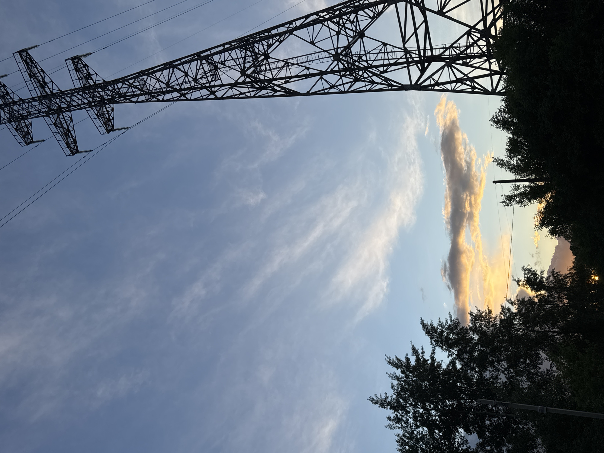 The photograph, taken in Chaikovsky, Russia, presents a low-angle view looking upwards. A black metal electricity pylon dominates the upper left portion of the frame. Electrical wires extend from the pylon. The sky occupies the majority of the image, revealing thin, wispy clouds. A notable vertical cloud formation, partially illuminated in golden light, appears on the right side of the sky. Silhouetted tree foliage fills the right edge of the frame and the lower right area. A power pole with wires is also visible amidst the foliage. The lighting suggests either a sunrise or sunset.