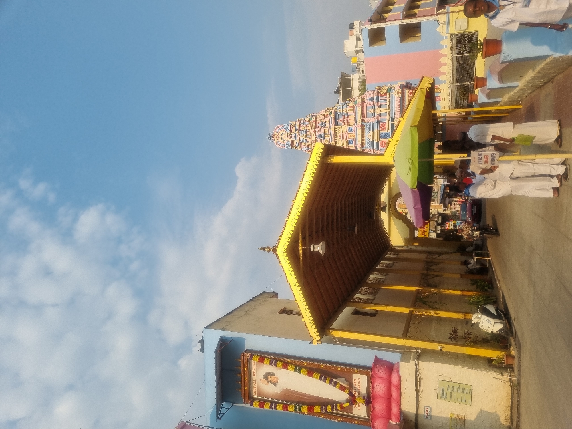 The image depicts a daytime street scene in Puttaparthi, India, likely an approach to a religious complex, with a prominent view of the Sathyabhama Temple gopuram.
On the left, a light blue building features a large, framed portrait of Sathya Sai Baba, adorned with colorful garlands, resting on a pink lotus-shaped base. Text above the portrait reads "LOVE ALL SERVE ALL," and below, "SAMASTHA LOKAH SUKHINO BHAVANTU." A "SILENCE 500FT" sign is mounted on the wall below. Extending from this side, a long, yellow-roofed covered walkway or archway with decorative trim runs into the midground. A white scooter is parked on the paved ground beneath this walkway.
In the center and right foreground, several adult males, predominantly dressed in white traditional Indian attire, are gathered under two large sun umbrellas (one purple and yellow, one green and yellow). They appear to be interacting. A white sign on a yellow pole reads "ENTRY STOP" and "DEVOTEES WITH LUGGAGE NOT PERMITTED." Behind these individuals, a multi-tiered, intricately sculpted temple gopuram, painted in vibrant hues of pink, blue, yellow, and red, rises prominently against the sky. To the right of the gopuram, a building with horizontal pink and light blue stripes is visible. In the far right foreground, an adult male in a white shirt and blue lanyard is partially visible.
The sky is clear blue with scattered white clouds, indicating fair weather. The scene is brightly lit by natural sunlight, casting visible shadows. A red car and a person on a dark motorbike are visible further down the covered walkway in the background. Potted plants line the base of the buildings and walkway walls.
