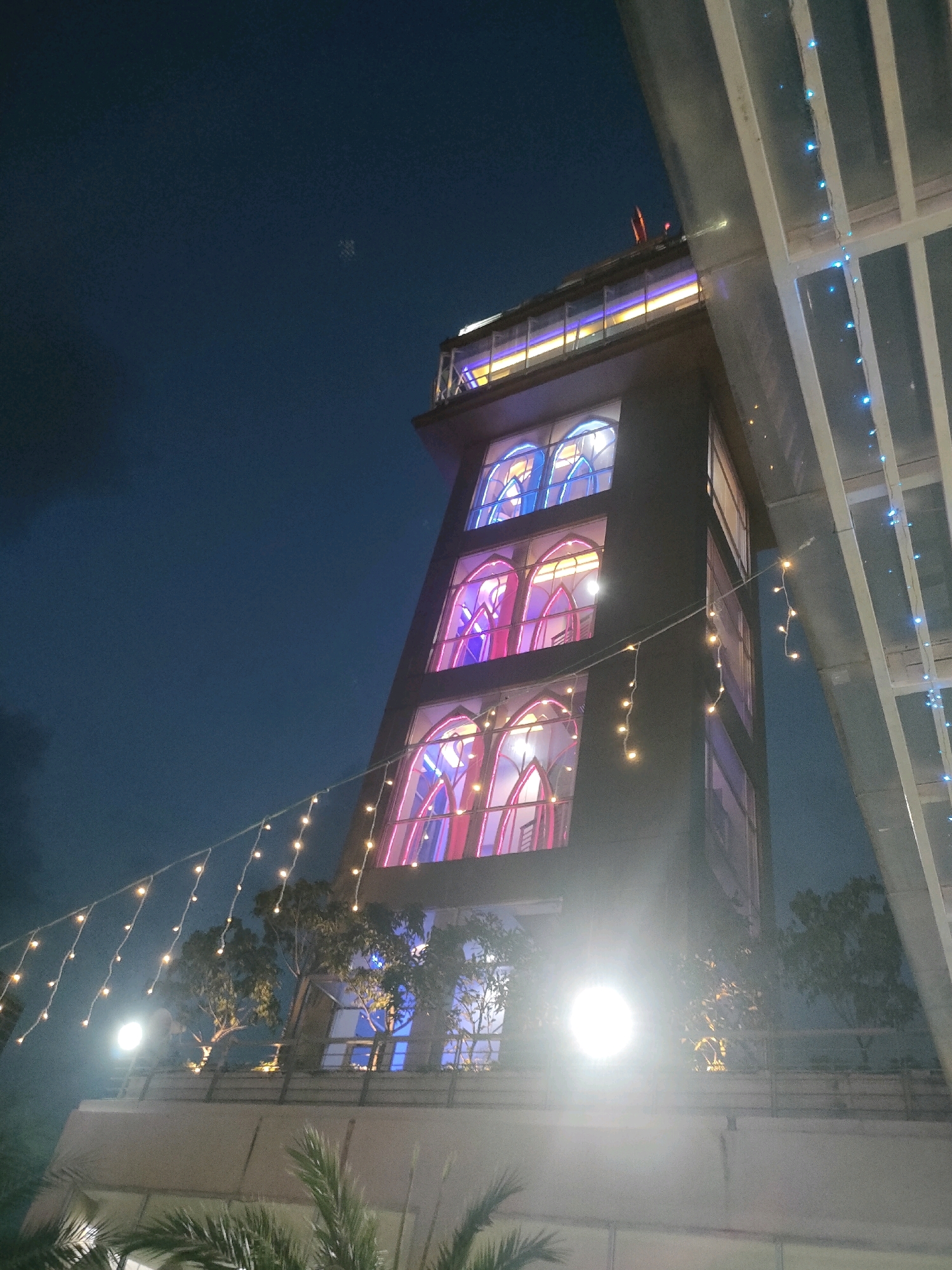 The image shows a modern building at night in Batam, Indonesia. The building is tall and dark, with illuminated arched windows that glow with pink and blue light. String lights, some yellow and some blue, are draped across the scene. Trees are visible on a lower level of the building, and palm fronds are in the foreground. The sky is dark blue, indicating it is nighttime. The overall impression is of a well-lit, contemporary structure against a night sky.