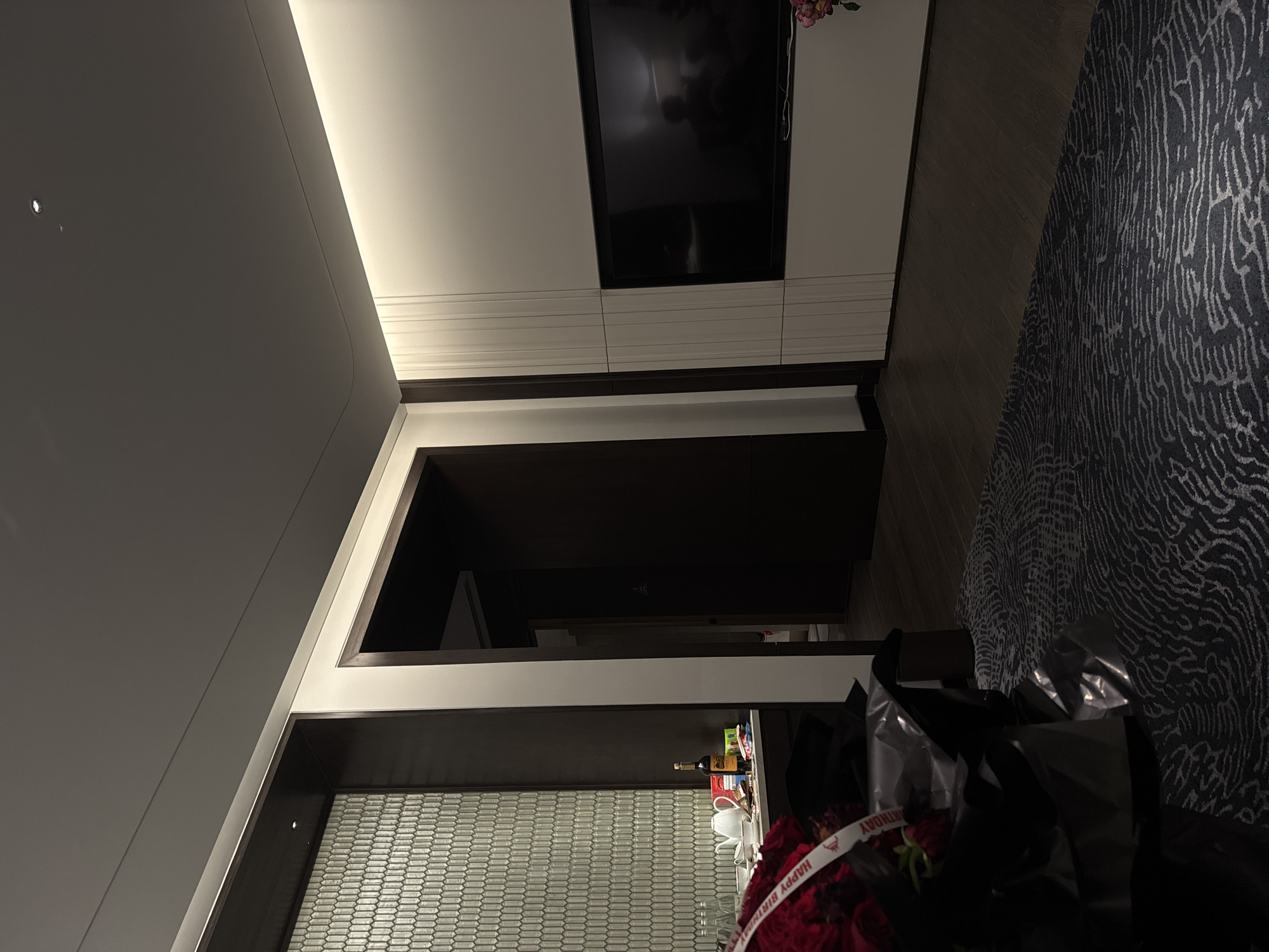 This is a slightly off-center, medium-angle shot of a hotel room interior. The room has modern design elements.
A flat-screen television is mounted on the wall, above a horizontal striped panel. To the right is a carpet with a dark, swirling pattern.
To the left, there's a glimpse into another part of the room, possibly a bathroom, through a doorway. A partial view of a counter is seen, with a bottle and other items placed on top of it. The lower portion of a privacy screen with a small, repetitive pattern is also visible.
In the foreground is a bouquet of red flowers in a black and silver bag with a "Happy Birthday" ribbon.