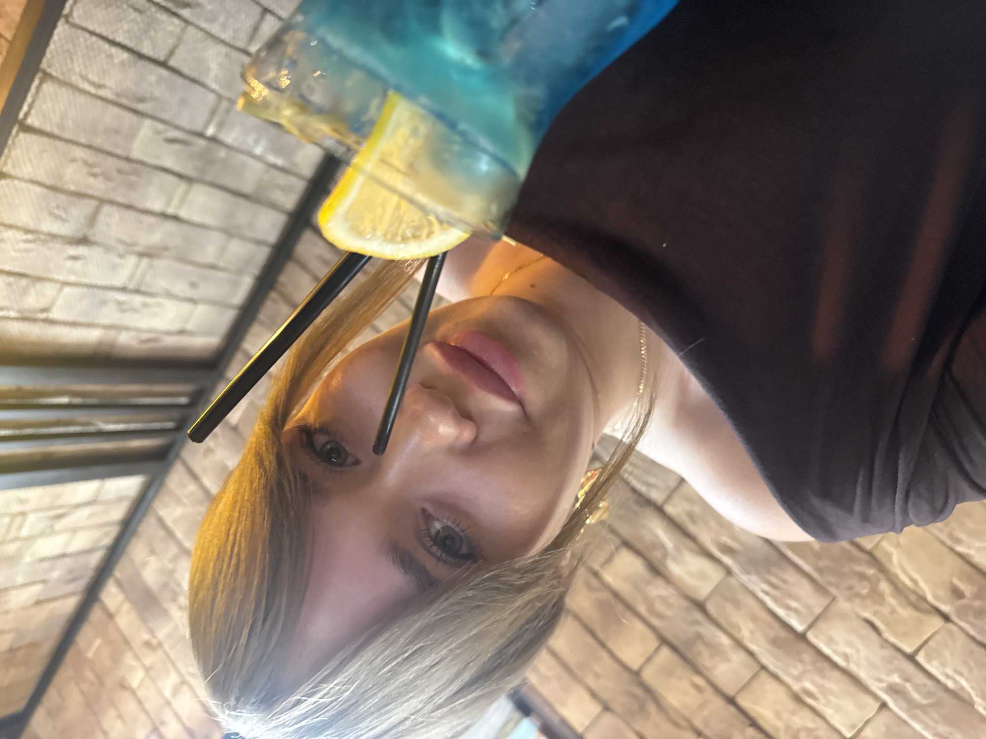 A young woman is holding a blue cocktail with a lemon slice and two black straws. She is wearing a black top and a gold necklace. She has blonde hair and is looking down at the camera. The background is a brick wall. It is not possible to tell if the location is in the city of Chaikovsky, Russia.