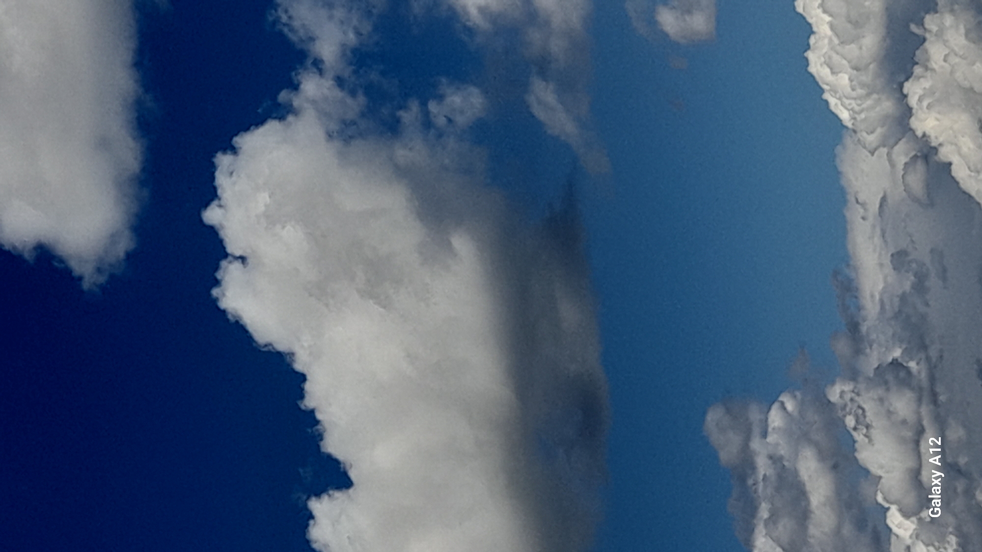 The image is a close-up, vertical shot of a bright blue sky filled with various types of clouds. The clouds range from fluffy white cumulus clouds, some casting shadows and appearing darker grey, to wispy cirrus clouds. The lighting suggests it is daytime, likely with the sun positioned to illuminate the clouds from above. In the bottom left corner of the image, the text "Galaxy A12" is visible, indicating it is a watermark from a Samsung mobile device. The scene depicts a natural outdoor environment, with no urban elements or people present. The location is stated as Марковский, Russia, but there are no visual cues within the image to confirm or deny this specific geographical context.