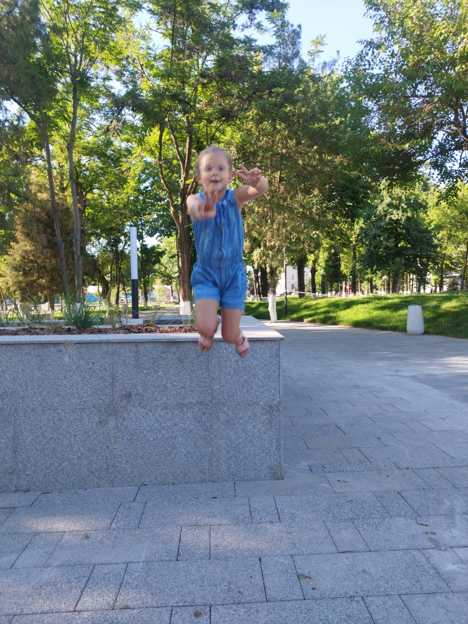 This media file captures a candid moment of a young girl in mid-air as she jumps over a stone wall in a park. The girl, wearing a denim jumpsuit and light pink sandals, has her arms raised, forming a peace sign with her fingers. Her expression is joyful and energetic, indicative of a playful activity.
The setting is a park with lush green trees and a clear blue sky, suggesting a bright, sunny day, likely in the late morning or early afternoon. The ground is paved with grey tiles, and a grey stone wall forms a prominent element in the foreground, with planters containing small bushes behind it. In the background, more trees and grassy areas are visible, along with a white lamp post and a light-colored trash receptacle.
The overall scene conveys a sense of childhood fun and freedom within a pleasant outdoor environment. No visible text is present in the image. The location is identified as Shymkent, Kazakhstan.
