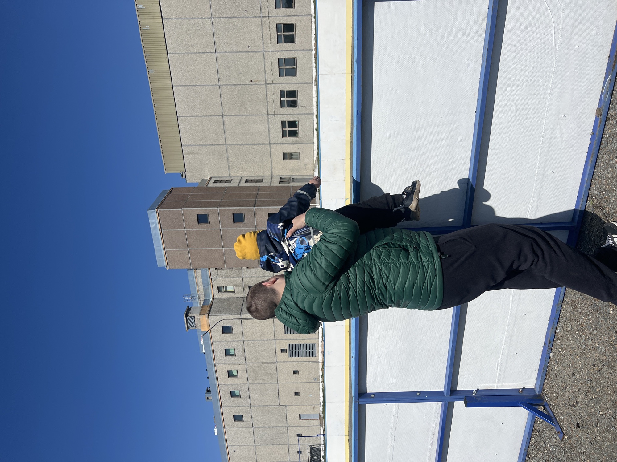 A man in a green puffer jacket and black pants is standing next to an ice rink with blue and white barriers. He is holding a young child in a blue jacket and yellow hat. The background includes a large, rectangular concrete building under a clear blue sky. The scene is in the city of Марковский, Russia.