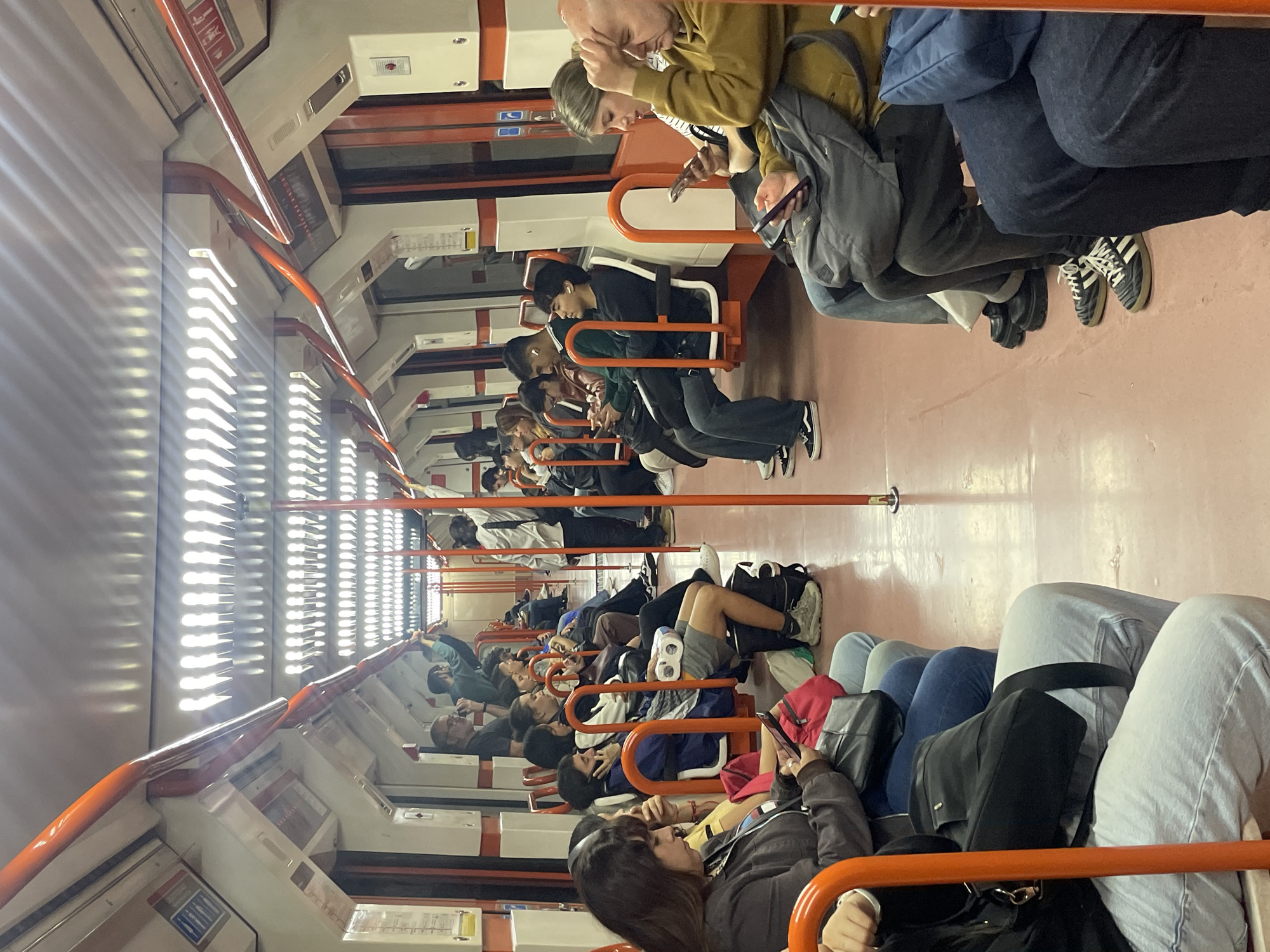 The image shows the interior of a Buenos Aires subway car. Passengers sit in rows of orange-framed seats, mostly looking at their phones. The car is well-lit, and the floor is a dull pinkish-tan color. The walls of the car are white, with orange accent lines. The car appears to be crowded.