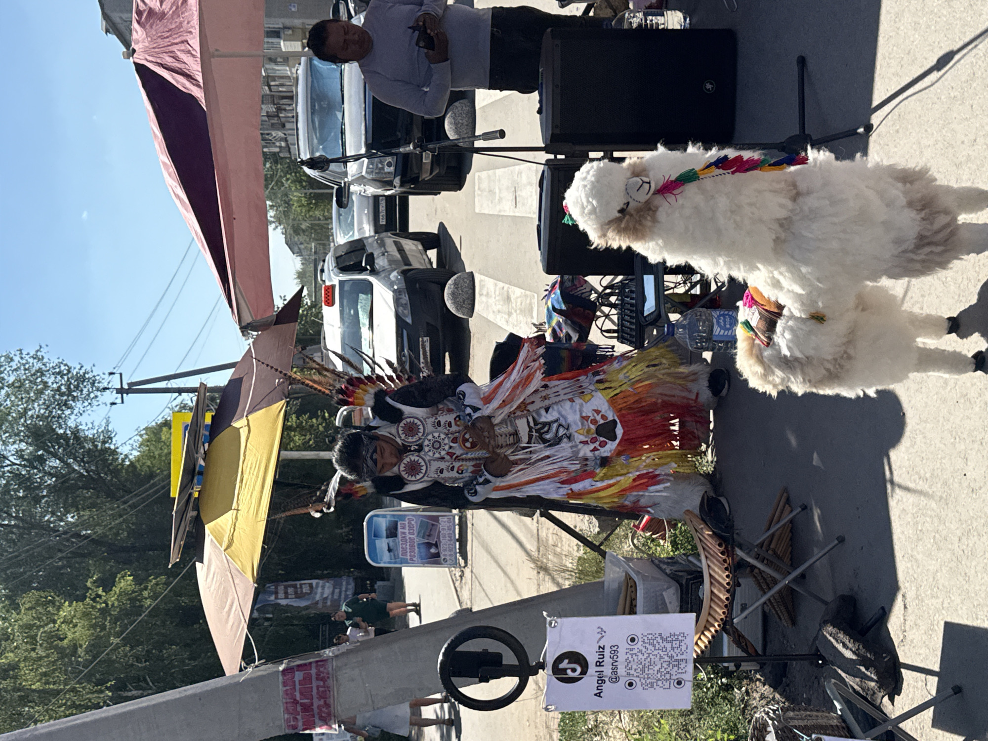 In Yarovoye, Russia, on a sunny day, a street performer dressed in traditional Indigenous attire, featuring a feathered headdress and a colorful fringed tunic, stands on a paved road. Beside them, a plush toy llama stands on a tripod, adorned with colorful tassels. The performer appears to be holding an instrument and looking down. In the background, several cars are parked along the street, and two large umbrellas, one yellow and one maroon, provide shade. A sign with a QR code and the username "@asrv593" is displayed on a stand near the performer. Other elements in the scene include speakers, a water bottle, and a foldable chair. The environment suggests an outdoor event or marketplace.