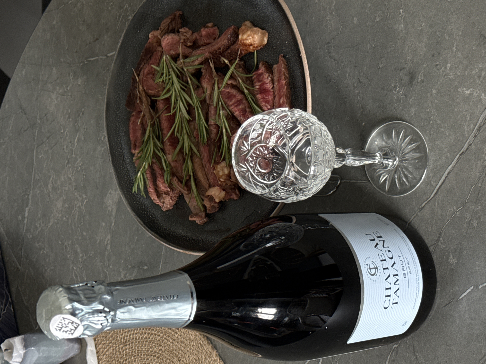 The image shows a close-up of a dark-colored plate containing sliced beef with rosemary sprigs on top, accompanied by an ornate crystal wine glass and a bottle of "Chateau Tamagne Brut" sparkling wine. The setting appears to be a tabletop with a grey, stone-like surface. There are no people visible. The environment doesn't offer clues to Krasnoyarsk, Russia.
