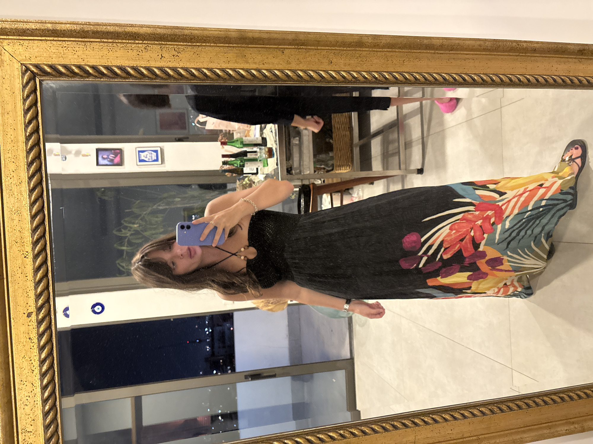 A woman is taking a mirror selfie in what appears to be an apartment with a balcony overlooking a city at night, likely Tel-Aviv, Israel. The woman is wearing a black strapless dress with a colorful floral print at the hem. She is holding a purple smartphone in her right hand, and her left hand is resting on her hip. The mirror is framed in a decorative gold frame. In the reflection, a second person can be seen in the background, standing near a table with bottles and glasses. The environment suggests a domestic setting, possibly a living room or dining area, with city lights visible through large windows in the background.