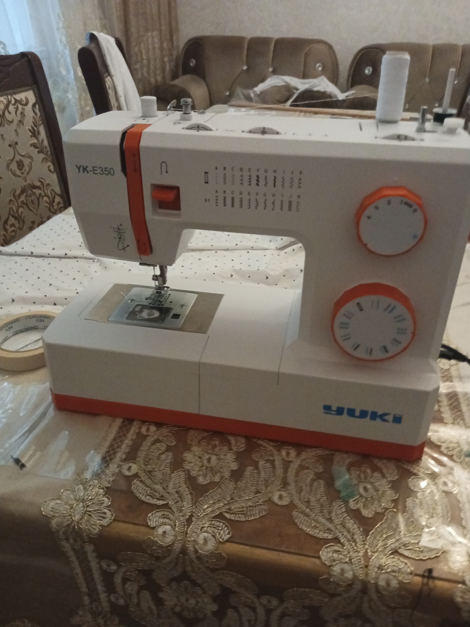 An indoor scene, likely a domestic setting in Masazir, Azerbaijan, captures a white and orange YUKI YK-E350 sewing machine positioned on a patterned tablecloth. The sewing machine is the central subject, with its needle and presser foot visible. A spool of white thread sits atop the machine. To the left of the sewing machine, a roll of masking tape is partially visible. In the background, blurred furniture, including a sofa and chairs, suggests a living or dining area. The lighting appears to be artificial, casting shadows and highlighting the textures of the tablecloth and furniture.