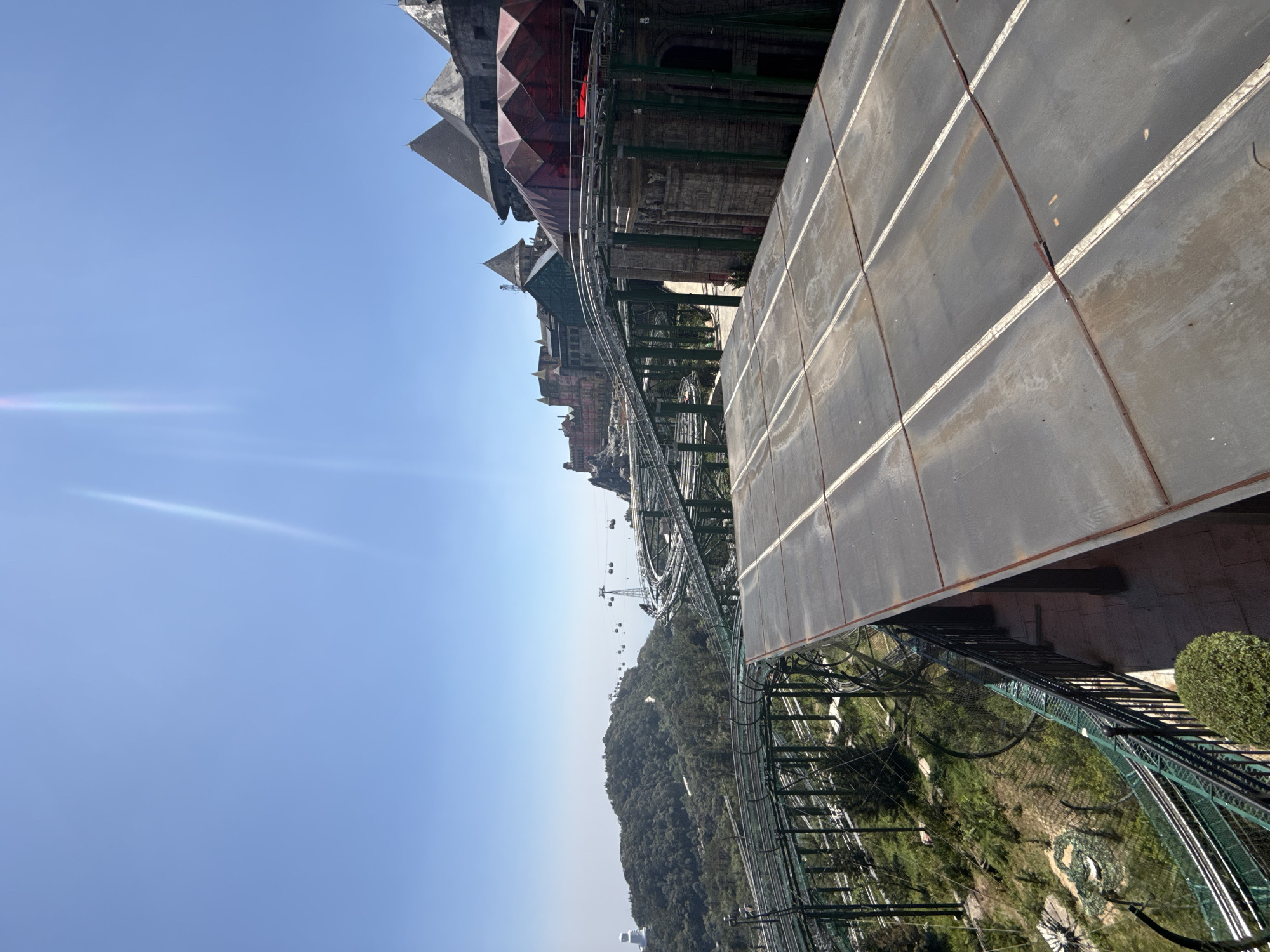 The image captures a scene at Ba Na Hills, a resort area in the city of Đà Nẵng, Vietnam. The perspective is elevated, possibly from a viewing platform or building.
Structures: Architectural elements reminiscent of European styles are visible, including a roof with red geometric panels and other building facades. A large, possibly metallic, structure with white lines runs along the right side of the frame.
Transportation: Cable car lines stretch across the landscape, with cable cars visible in the distance. A winding track system, possibly for a roller coaster or similar ride, is prominent.
Environment: Below the tracks and cable cars is a landscape covered with dense green vegetation.
Atmosphere: The sky is clear and blue, with a lens flare effect present in the upper left portion of the frame.
No people are explicitly visible in the image.