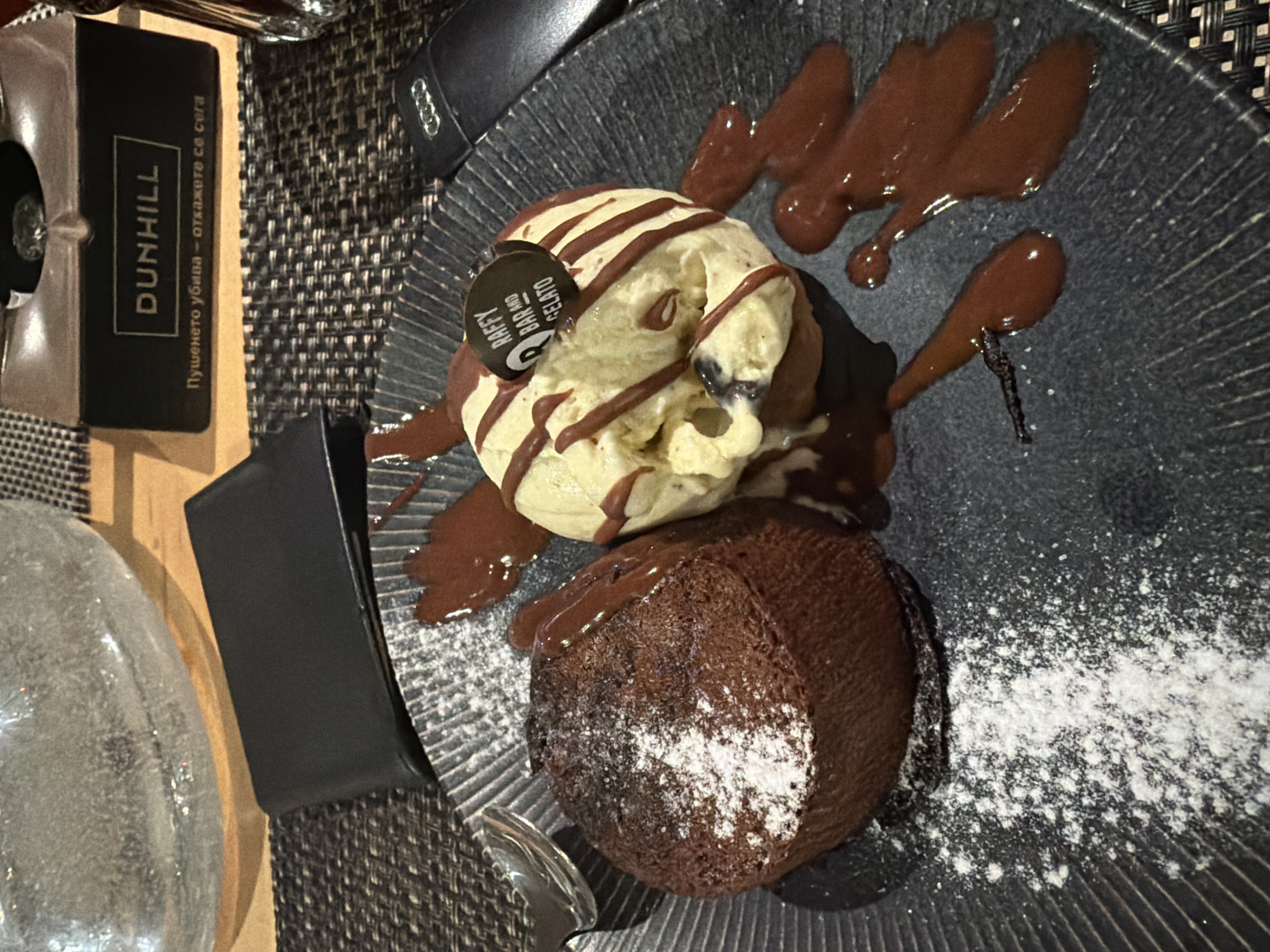 The image shows a dessert plate with a chocolate lava cake dusted with powdered sugar and a scoop of ice cream drizzled with chocolate syrup. The plate is dark and textured. Next to the plate, there is an ashtray with the Dunhill brand name, a spoon, a glass, and a black object. The photo was most likely taken in a restaurant, possibly in Plovdiv, Bulgaria, given the Bulgarian text on the ashtray. The Raffy Bar and Gelato logo is visible on a small sign near the ice cream.
