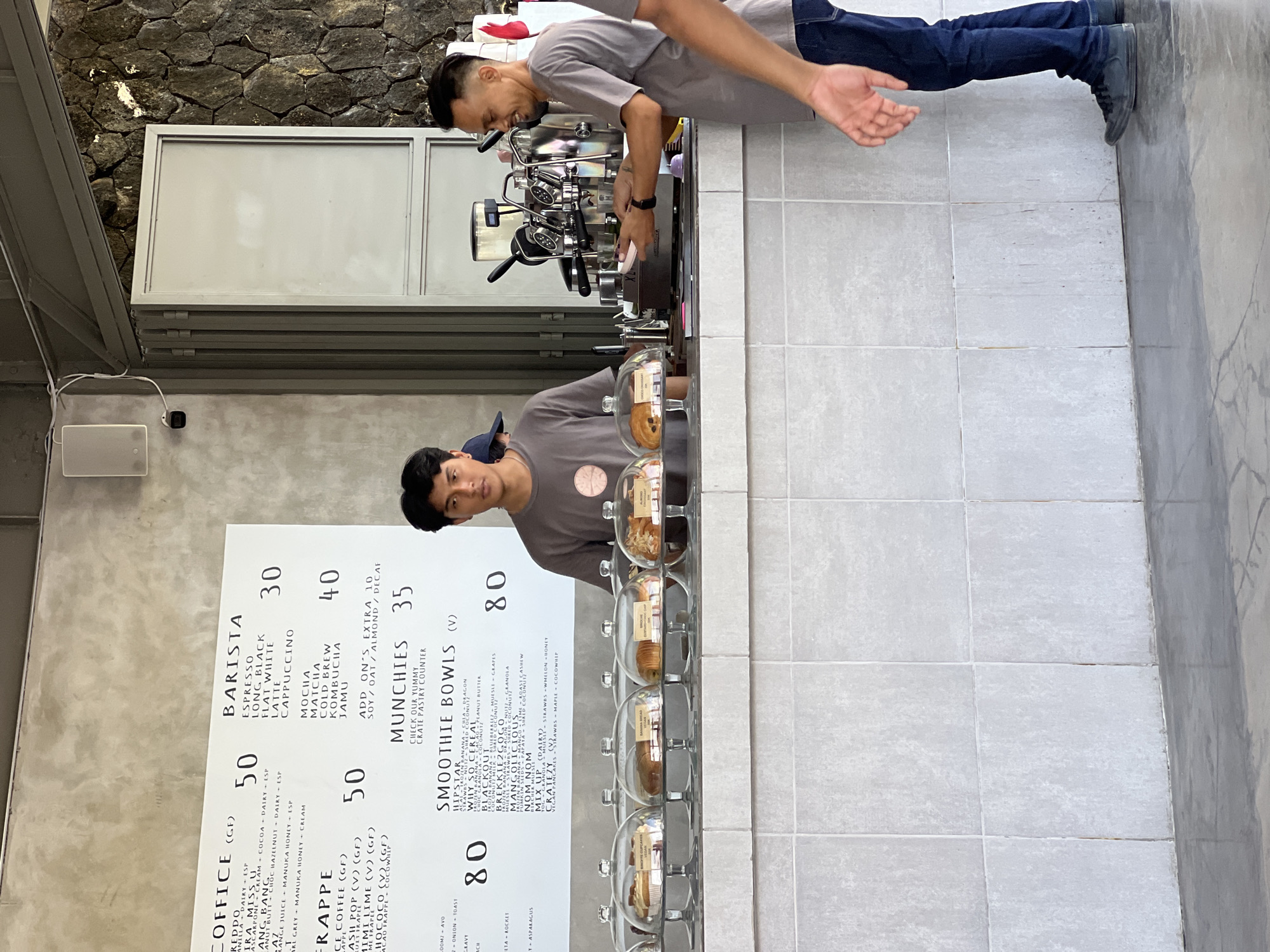 The image shows the interior of a cafe in Mengwi, Indonesia, with a menu displayed on a concrete wall. Two men are visible behind a counter where pastries are displayed under glass domes. The menu lists various coffee drinks, frappes, smoothie bowls, and "munchies." One man, wearing a grey t-shirt and jeans, is operating an espresso machine. The other man, also in a grey t-shirt, is standing near the pastries. The cafe appears to have a modern, industrial aesthetic with concrete walls and stone accents. The overall atmosphere is that of a busy cafe with staff preparing orders.
