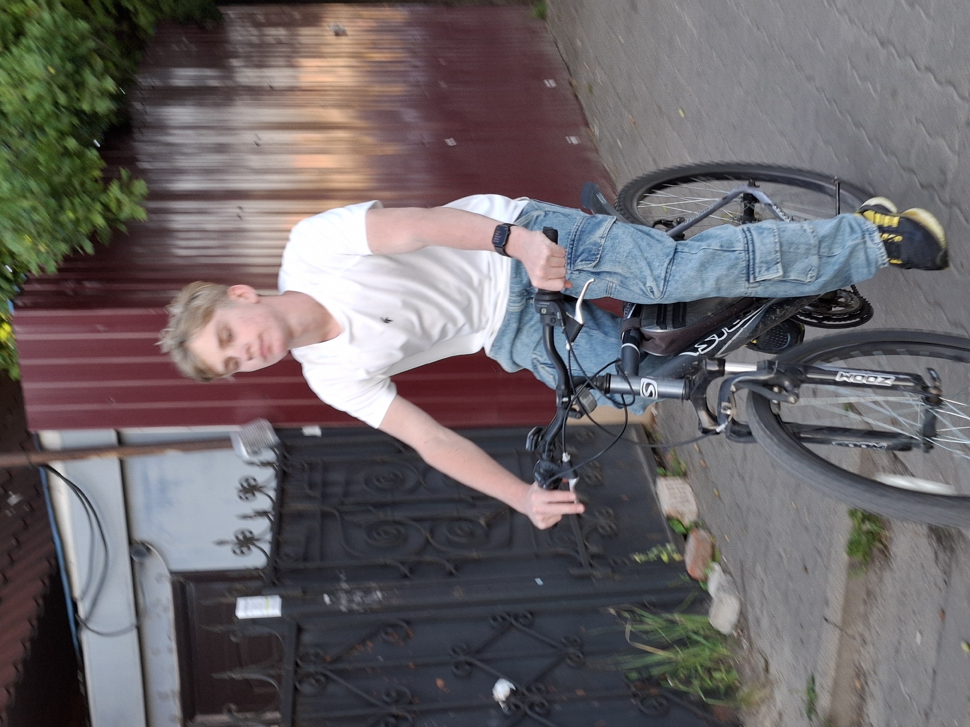 A young, blond-haired male wearing a white t-shirt and light blue cargo pants rides a black and silver bicycle down a paved path next to a dark metal fence. He is looking downwards. The bicycle has a black seat, black handlebars with black grips, and a black water bottle cage mounted beneath the handlebars. The front wheel of the bicycle has white spokes. A black digital watch is worn on his left wrist. Behind the fence is a reddish-brown corrugated metal structure. Trees with green leaves are visible above the metal structure. The ground is gray asphalt with some patches of green grass growing through cracks. The scene appears to be outdoors during daylight.
