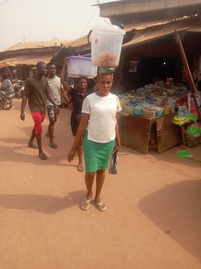 A woman carries a clear bucket full of brown objects on her head in an outdoor market. She wears a white shirt, green skirt, and sandals, and holds a bag. Behind her, a woman balances a purple container on her head, followed by two men. They walk along a dirt road lined with shops and stalls. A variety of goods are visible in the shops, including baskets and containers. The scene is brightly lit, suggesting a sunny day.