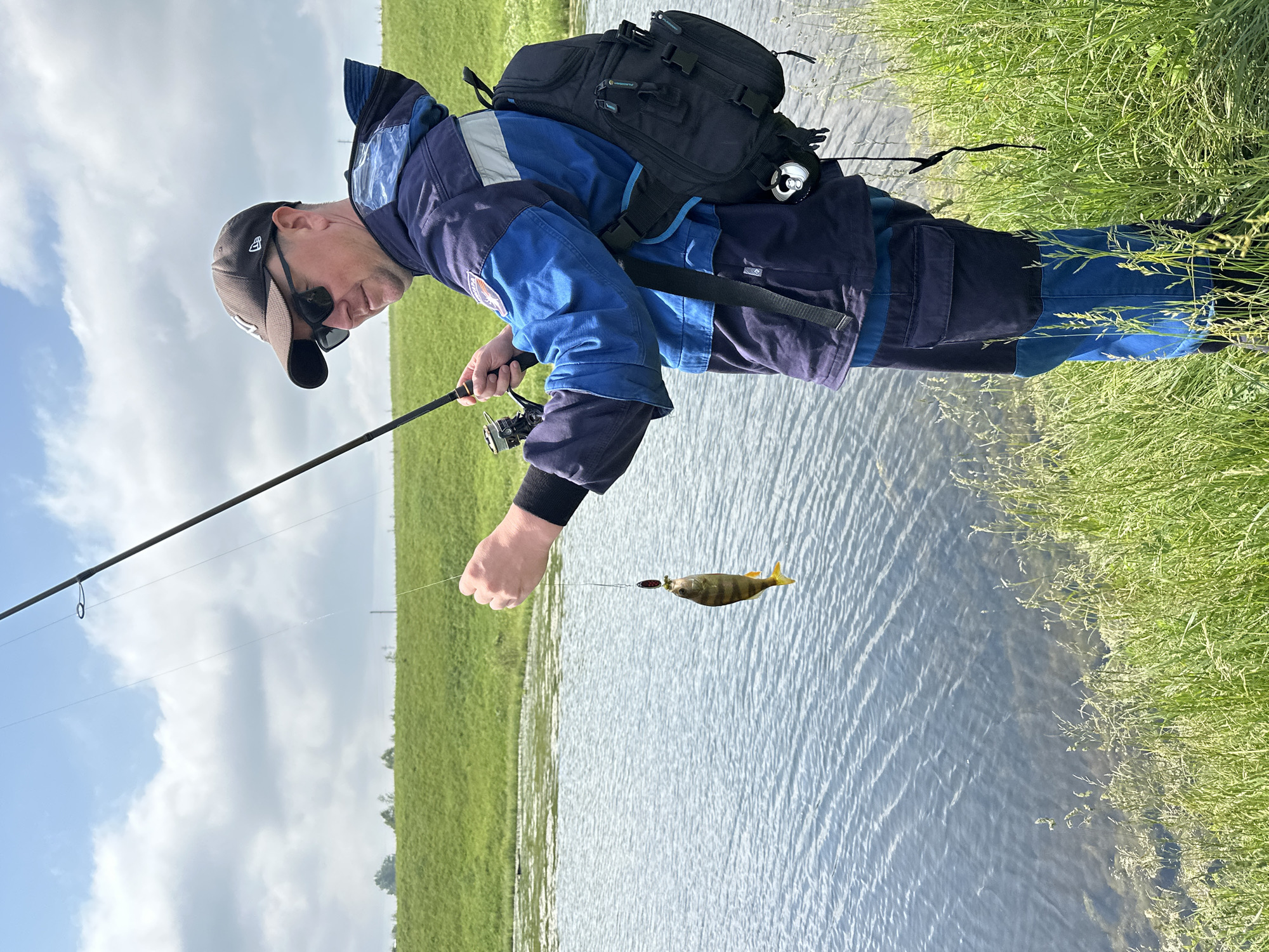 A man stands on the grassy bank of a body of water, holding a fishing rod with a small fish dangling from the line. He is wearing a blue and gray jacket, dark pants, a baseball cap, and sunglasses. A black backpack is slung over his shoulder. The water is calm with visible ripples, and the background features a green, grassy hill under a cloudy sky. The scene appears to be a casual outdoor activity.