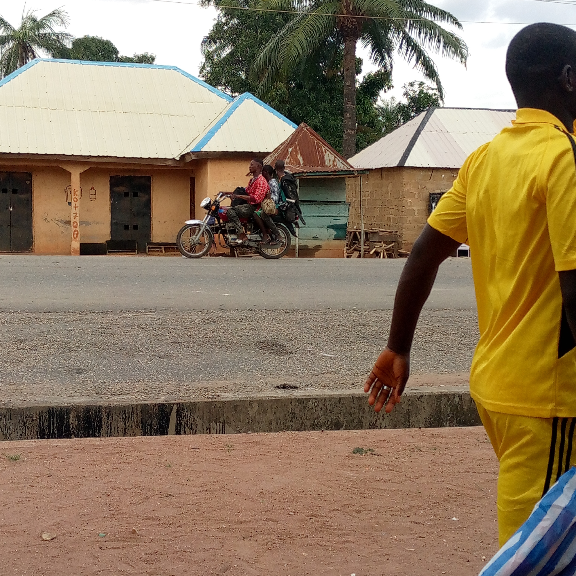 The image depicts a street scene in what appears to be Mararaba, Nigeria. In the background, three people are riding on a motorcycle along a paved road. The buildings are simple, single-story structures with tin roofs. A palm tree is visible in the background. In the foreground, a man wearing a yellow tracksuit is walking away from the camera. The ground is reddish dirt with a concrete curb separating it from the paved road.