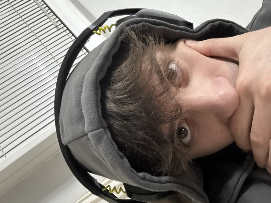 The scene depicts a close-up portrait of a young adult male indoors. The provided location of Selanovtsi, Bulgaria, is not visually confirmed by elements within the image itself. The individual, with light skin and brown wavy hair partially visible under a raised grey hooded sweatshirt, is wearing black over-ear headphones with yellow coiled cables visible on each side. The subject's brown eyes maintain direct contact with the camera. Their left hand is positioned with fingers pressed against the lower jaw and mouth, partially obscuring the mouth area. The background primarily consists of light-colored walls or ceiling, with a white, louvered ventilation grate or lighting fixture visible overhead in the upper portion of the frame.