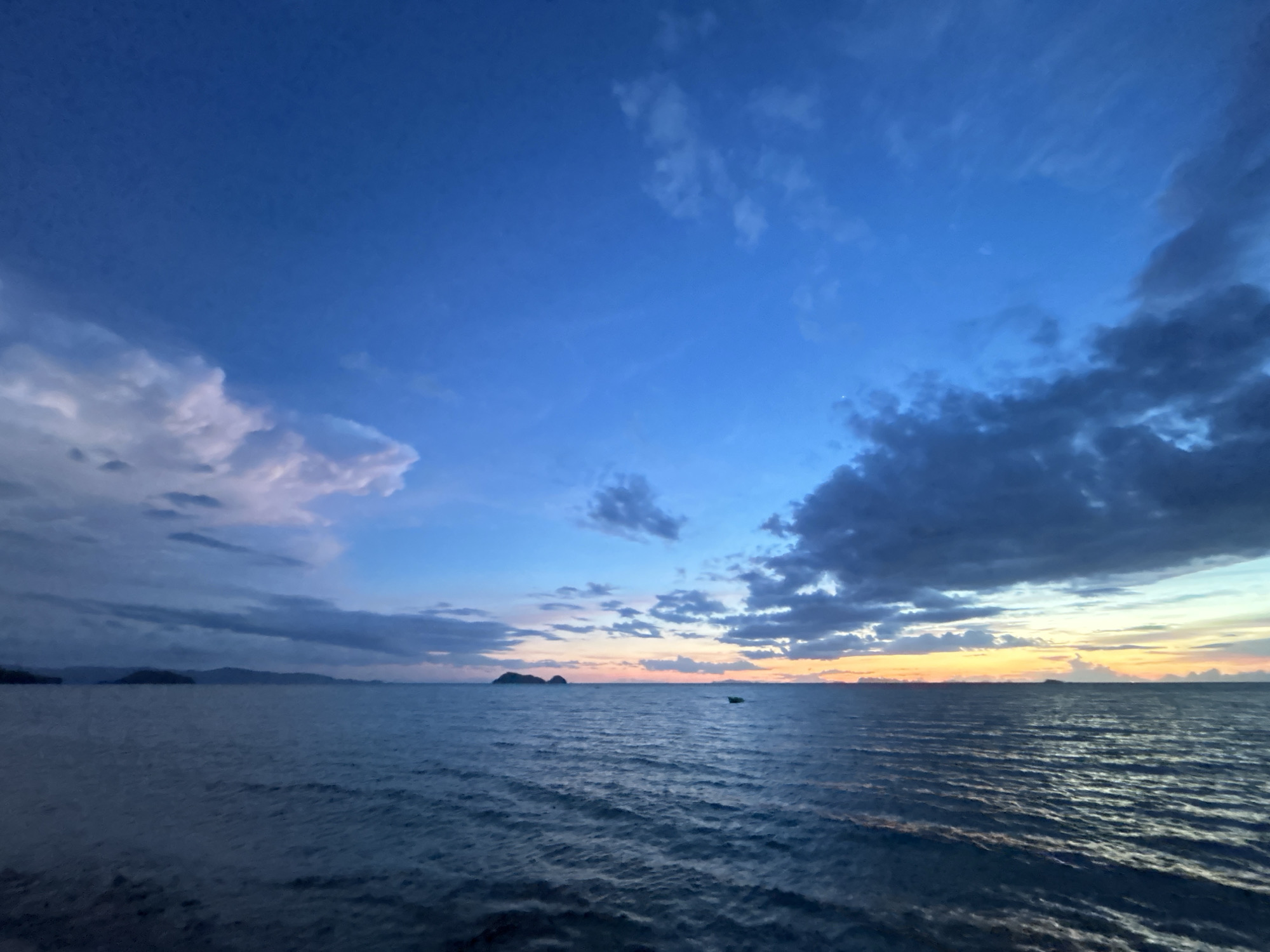 The image depicts a serene seascape at dusk in the Ko Pha-ngan District of Thailand. The sky is a gradient of deep blues, fading into lighter hues near the horizon, where a band of soft orange and yellow signals the setting sun. Fluffy clouds, tinged with pink and purple, are scattered across the sky, contrasting with darker, more ominous clouds on the right. The calm sea, a deep blue, reflects the twilight sky. In the distance, the silhouettes of islands or a mainland coastline are visible, appearing as dark, low-lying shapes against the horizon. A small, dark boat is present on the water, positioned to the right of the center. The overall impression is one of tranquility and natural beauty.
