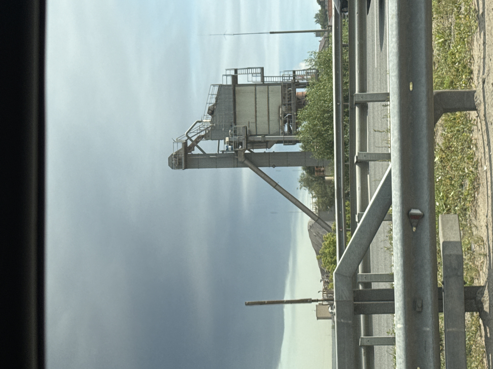 The image shows a large industrial structure, likely an asphalt or concrete plant, located near a highway in Domodedovo, Russia. The plant consists of a tall, multi-level metal tower with a series of conveyors, walkways, and enclosed sections. A large, sloping conveyor belt extends from the ground to the upper levels of the tower. In the background, there is a pile of aggregate material, green vegetation, and a dark, ominous cloud formation in the sky. The foreground features a metal highway guardrail and roadside grass. The scene appears to be captured from a moving vehicle, indicated by the blur in the upper portion of the image and the framing of the shot. No people are visible in the image.