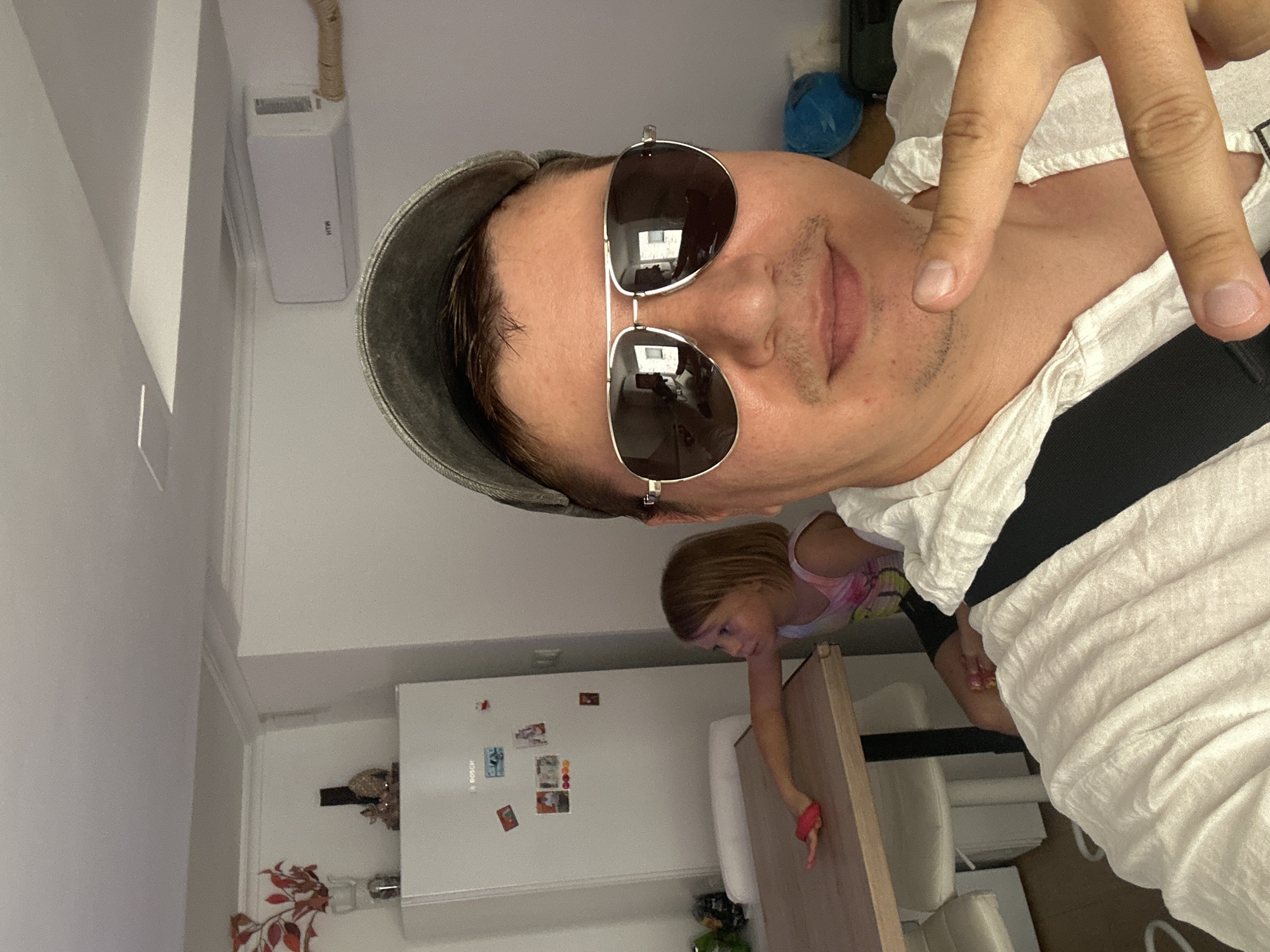In an indoor scene, a man wearing sunglasses and a cap takes a selfie. To his left, a young girl sits at a wooden table, her arm extended with a pink object in her hand. Behind the girl, a white refrigerator is visible, adorned with several magnets. The room appears to be a kitchen or dining area, with a portion of an air conditioning unit mounted high on the wall. The overall impression is of a casual, domestic setting. The image does not provide any specific visual cues to place it in Benimàmet-beniferri, Spain.