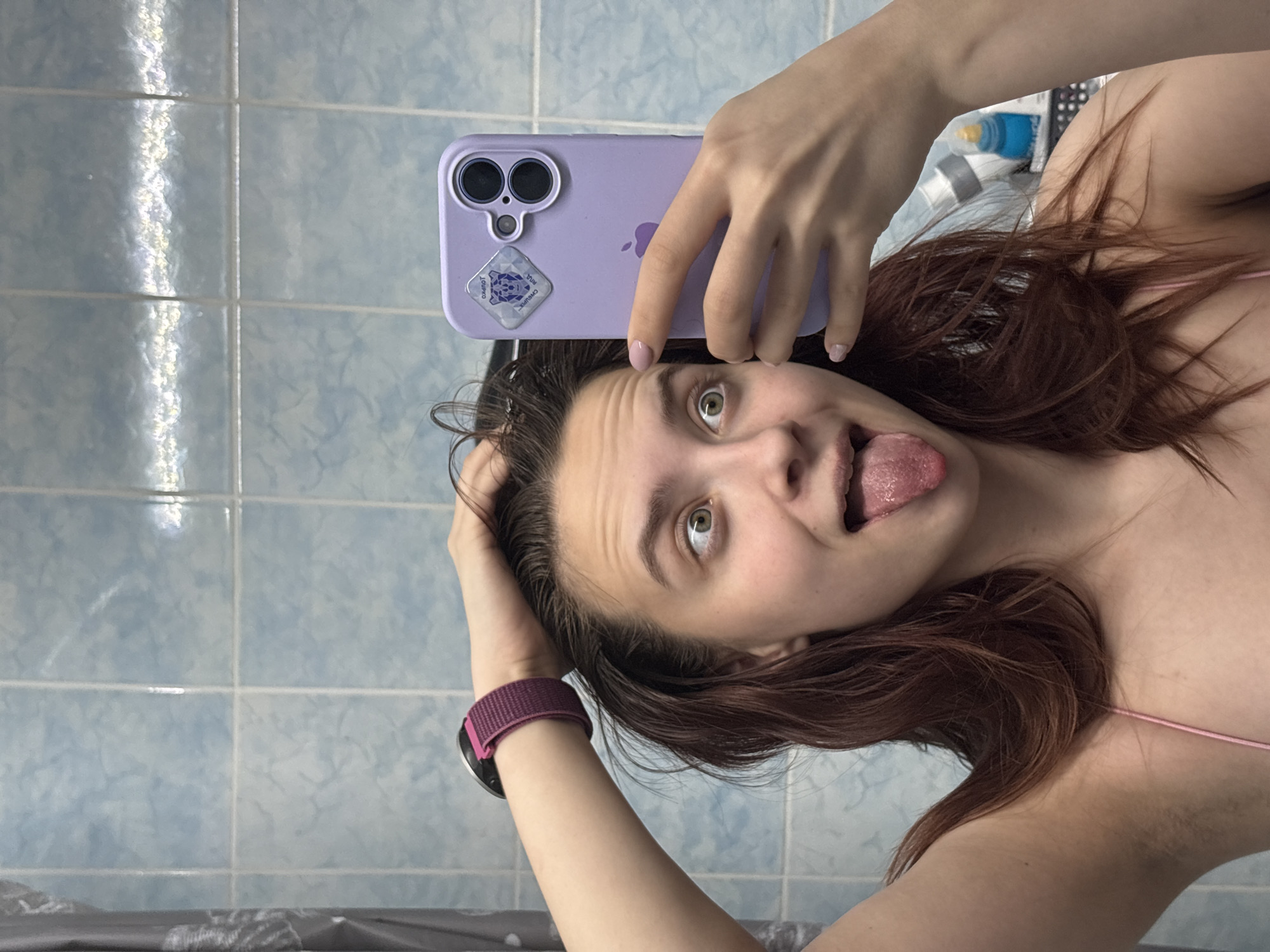 A woman is taking a selfie in a bathroom. She is holding a purple iPhone in her right hand and has her left hand on her head. She is sticking her tongue out and looking at the camera. She is wearing a pink top and a purple watch. The bathroom has blue and white tiles. She is possibly in Прикамский, Russia.