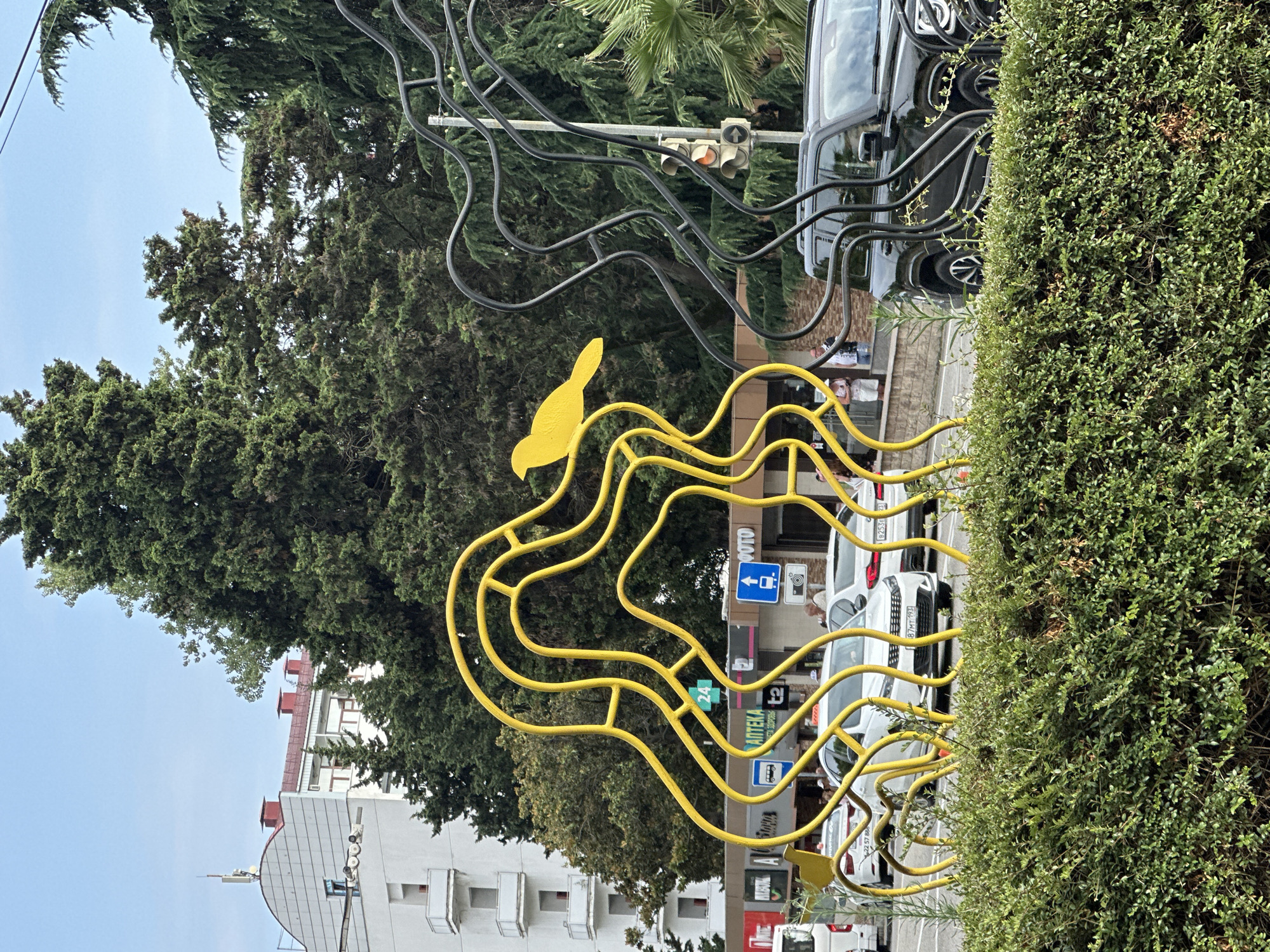 A yellow sculpture of a bird perched on swirling branches is in the foreground of the image, with a black sculpture of similar design to its right. Behind the sculptures is a street with cars and pedestrians. Buildings and tall trees are visible in the background. The scene appears to be a city street in Sochi, Russia, likely in a park or public square.