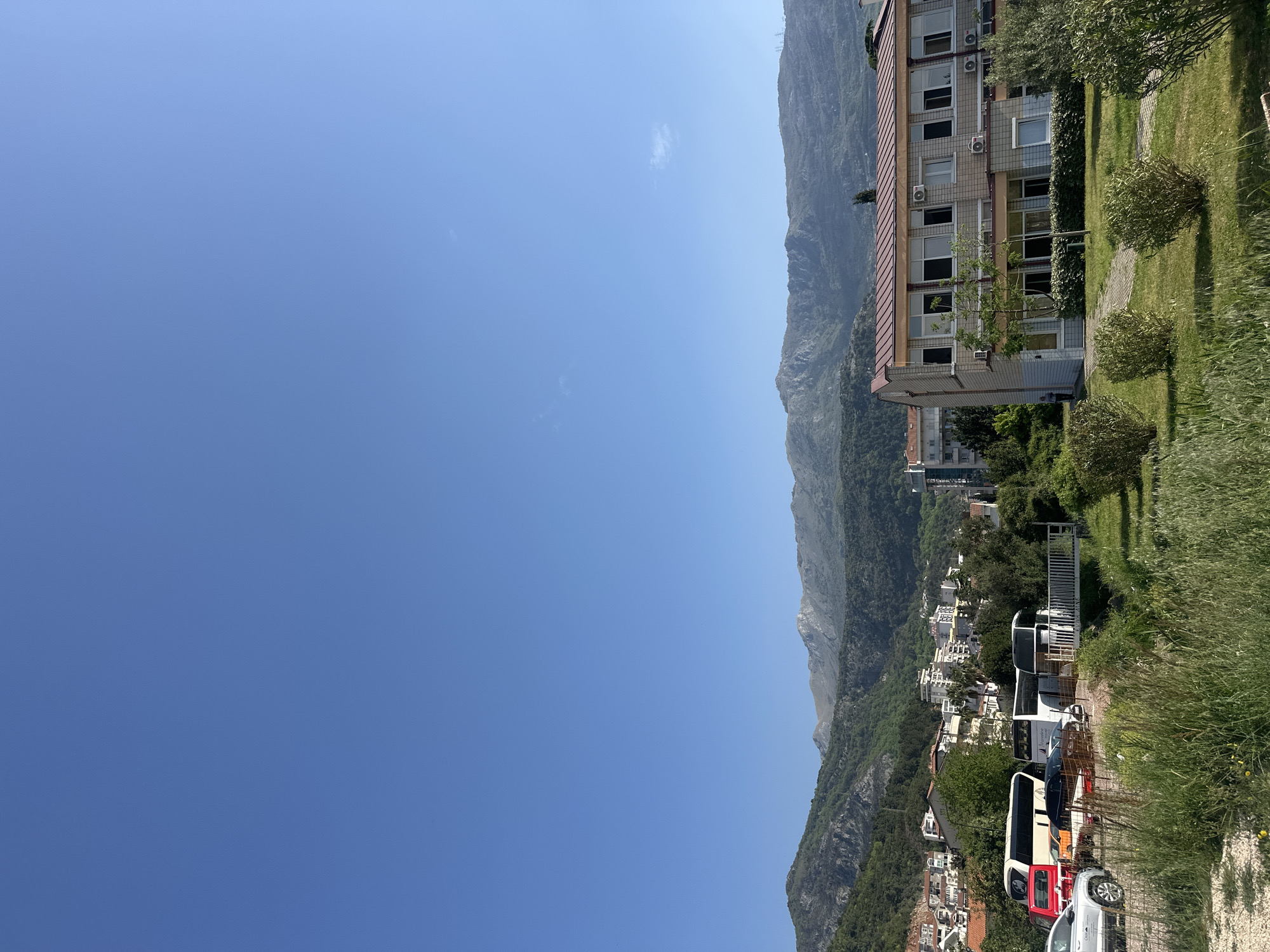 This image presents a serene landscape captured under a vast, clear blue sky, indicative of a bright, sunny day, likely late morning or early afternoon. Only a few faint, wispy clouds are visible in the expansive sky that dominates the upper left portion of the frame.
A majestic range of green, tree-covered mountains stretches across the mid-ground, forming a rugged and picturesque backdrop. At the base of these mountains, on the right side of the image, lies a populated area that appears to be part of a town or village, consistent with locations like Budva, Montenegro.
In the immediate right foreground, a multi-story building with a light-colored facade and a reddish-brown tiled roof stands out. It features numerous windows and several visible air conditioning units. A lush green lawn with scattered bushes and a paved path lies in front of this building. Further down the mountain slope, more light-colored residential or commercial buildings are nestled among trees.
The lower right foreground reveals a parking area or roadside where several vehicles are parked. These include a large white tour bus, a smaller dark-colored van or bus, a red truck, and a silver car. White metal fencing and tall grasses are also visible in this area.
No specific human activity is taking place; the scene is a static depiction of the natural and built environment. The bright light and clear visibility suggest pleasant weather conditions. The overall impression is one of tranquility in a scenic, mountainous region, reflecting the typical landscape found in coastal towns such as Budva, Montenegro. No discernable text is clearly visible on any objects within the image.