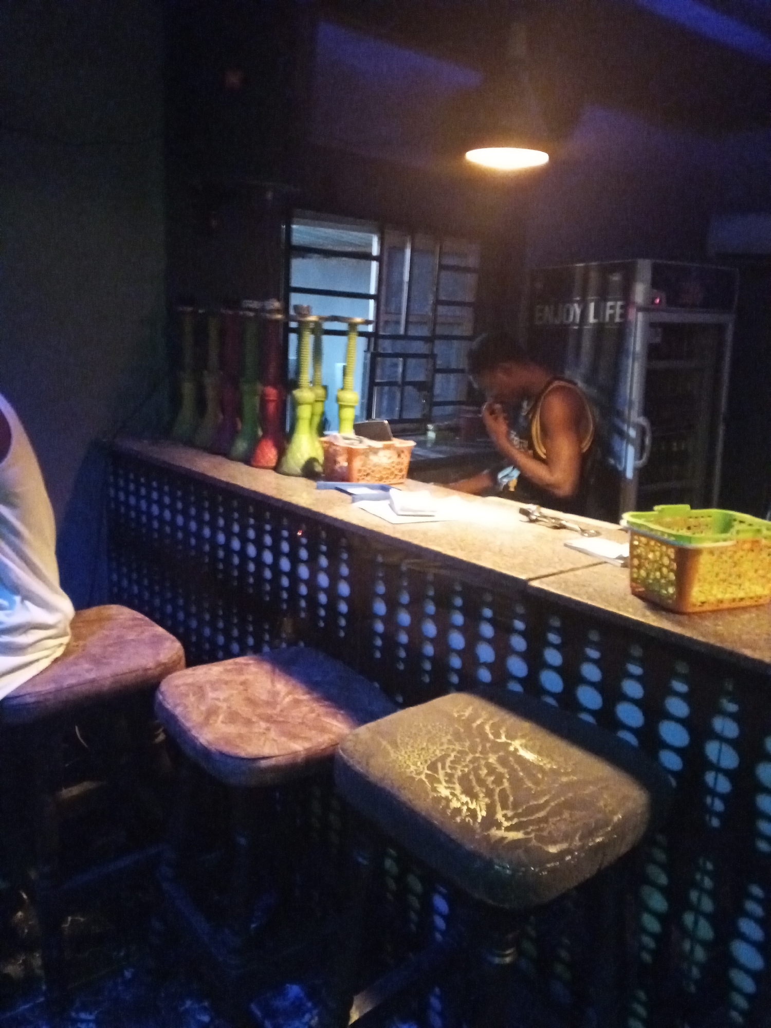 The image shows the interior of a dimly lit bar in Lokoja, Nigeria. A bar counter with a perforated metal facade dominates the scene, with three bar stools positioned in front. Various hookahs are lined up on the counter. A man wearing a tank top stands behind the counter, seemingly on his phone. A refrigerator labeled "ENJOY LIFE" is visible behind the man. A single light fixture illuminates the scene. Several baskets and a pair of tongs are on the counter. The floor is dark and indistinct.
