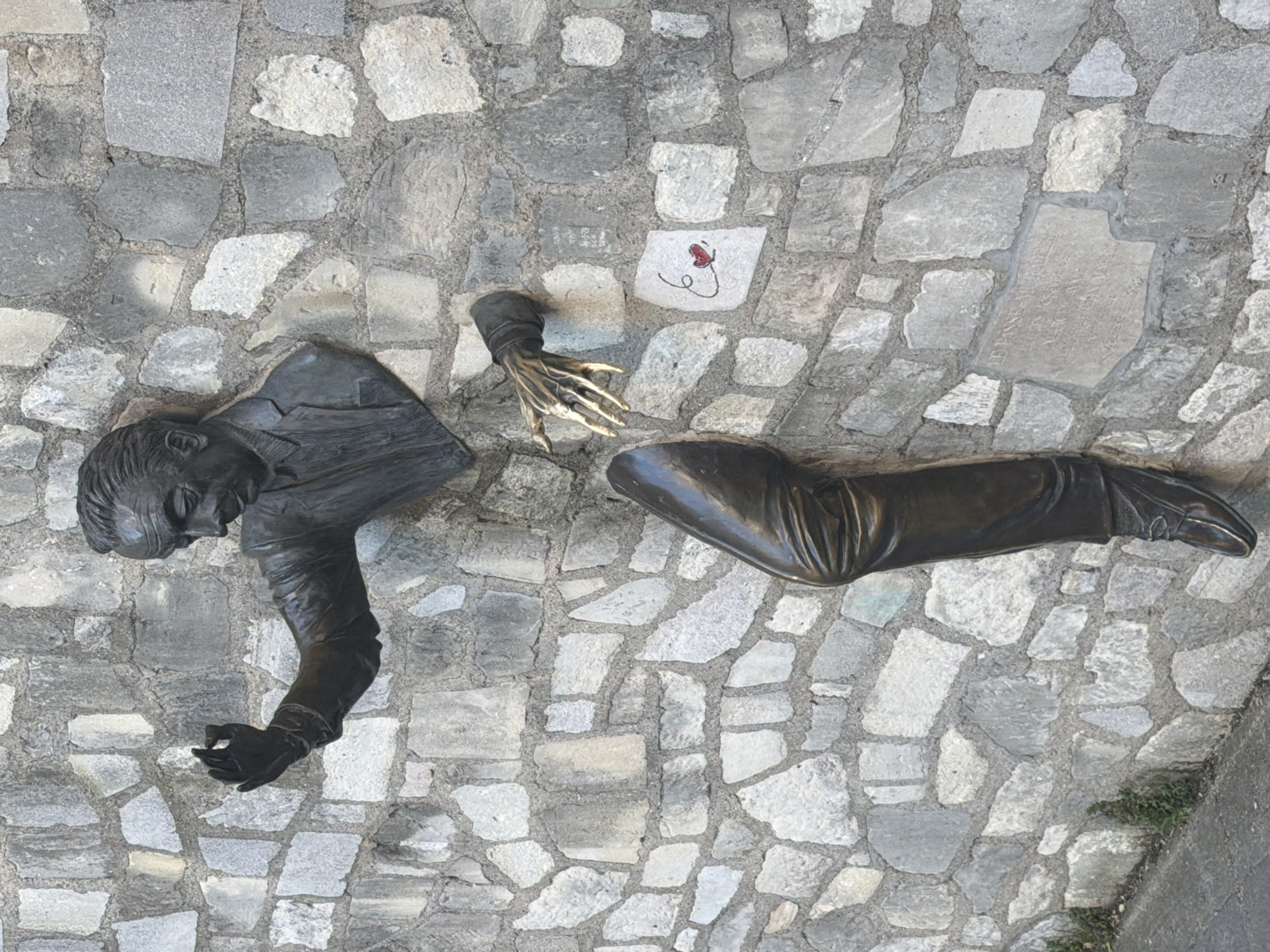 This image shows a sculpture of a man seemingly emerging from the cobblestone pavement. The sculpture is bronze-colored and depicts the man's head, upper torso with an arm reaching out, a skeletal hand, and legs. The sculpture appears integrated into the cobblestone ground, which is made of gray stones. There is a small drawing on one of the stones near the hand. The location is likely Paris, France, as the sculpture is a well-known piece in the Montmartre district.