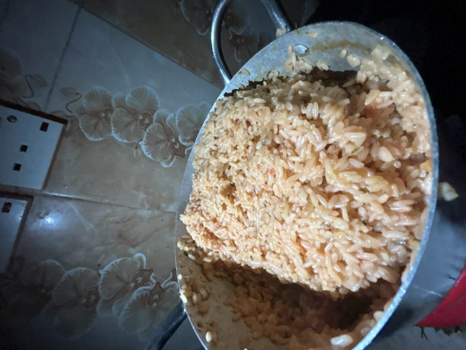 This image captures a large, silver-colored metal pot brimming with reddish-orange cooked rice, consistent with jollof rice, set within an indoor environment in Egbe, Nigeria. The rice appears thoroughly cooked, with individual grains visible along with hints of seasonings or vegetables. The pot, full to its rim, shows one metallic handle on the right side. The background consists of a tiled wall featuring a light brown base with a repeating pattern of white or light-colored orchid-like flowers. Two electrical outlets or switches are visible on the upper part of the tiled wall. No individuals are present in the scene, and no specific actions are depicted beyond the static presentation of the cooked meal.