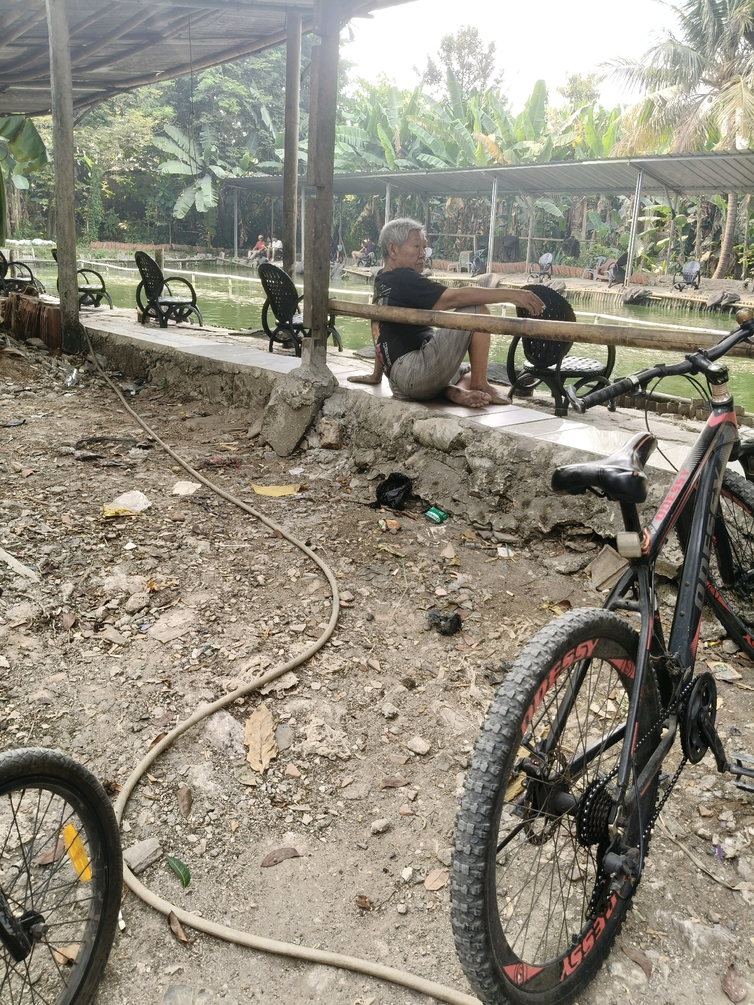 In the city of South Tangerang, Indonesia, this image depicts an outdoor scene adjacent to a body of water. In the foreground, a bicycle with a black frame and red accents is parked. Its knobby tires suggest it is a mountain bike. Behind the bicycle, a tan-colored hose lies on the ground, which is covered in dirt, rocks, and fallen leaves. A concrete barrier runs along the edge of the water, and on top of this barrier, an elderly man with gray hair is seated, wearing a black t-shirt and checkered shorts. He is looking to his left, with his hands resting on a bamboo railing. Several black wicker chairs are visible behind him, arranged along the edge of the water. The background is filled with lush green foliage, including banana trees and palm trees, under a partially covered shelter. Other people can be seen in the distance, seated or standing near the water's edge. The overall environment suggests a recreational area or park.