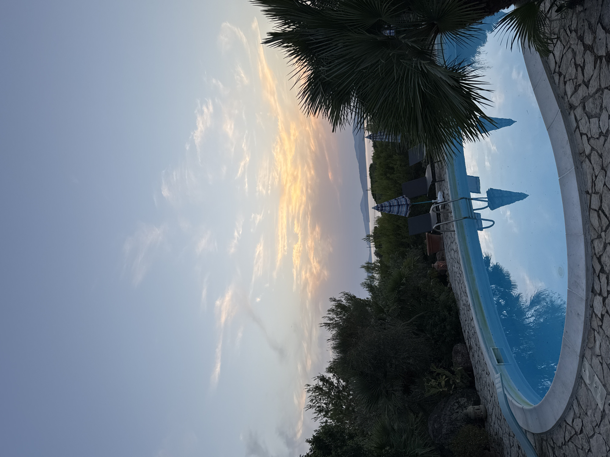 A wide-angle, eye-level shot captures an outdoor swimming pool at dusk. The pool, with a stone border and surrounding paved area, is oval-shaped and filled with clear blue water. The water reflects the sky and surrounding vegetation. Three lounge chairs with umbrellas are positioned by the pool. Lush green foliage and a palm tree frame the scene on the right. In the background, a body of water separates the foreground from distant hills and islands. The sky is a blend of pale blue and soft orange hues, with wispy clouds indicating the setting sun. The location is in Alghero, Italy.