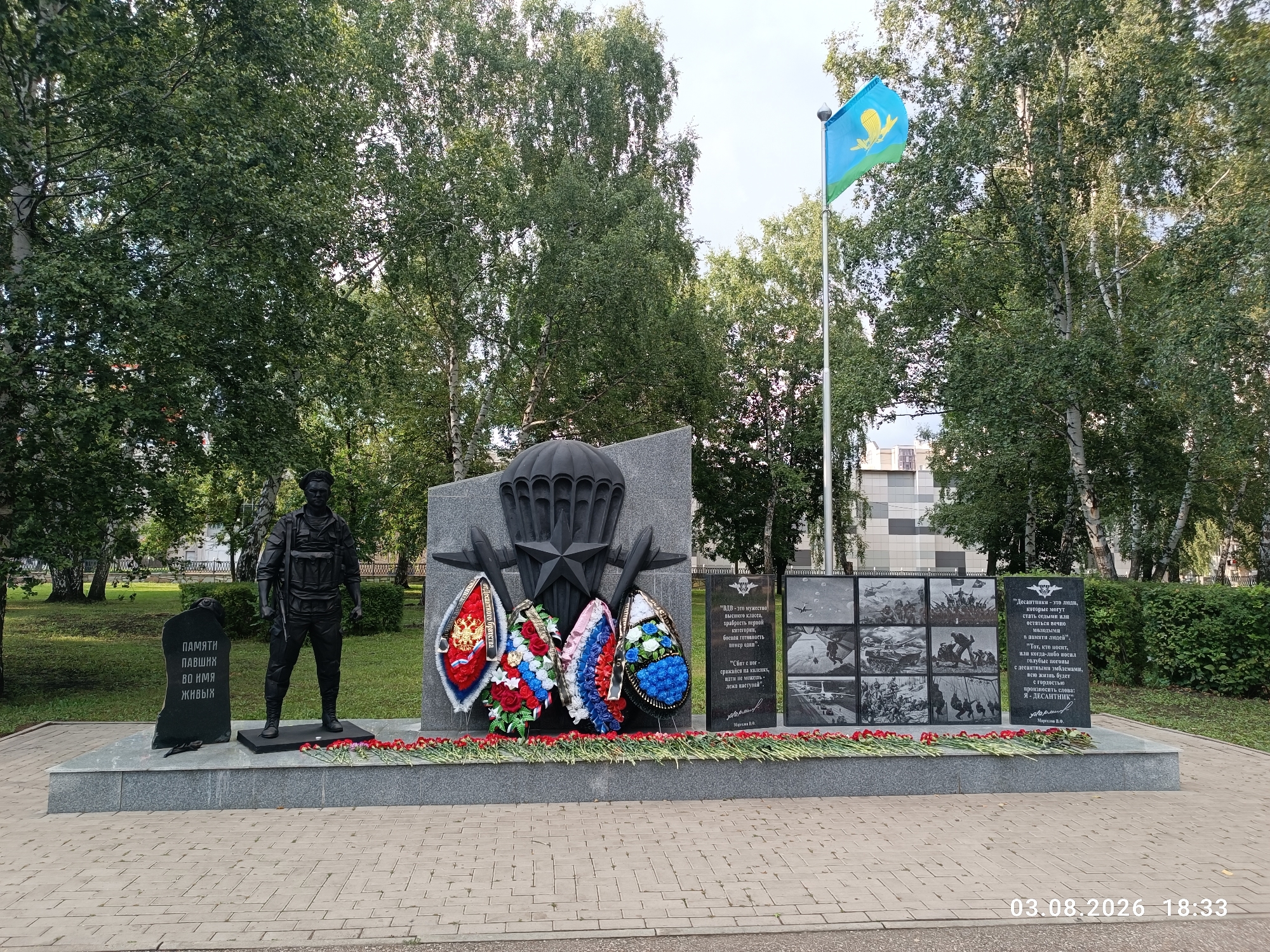 The image shows a monument dedicated to paratroopers in Sterlitamak, Russia. The monument features a statue of a paratrooper, a large granite structure with a parachute emblem, and several wreaths. A flag pole with a flag is also visible to the right of the monument. The monument is surrounded by trees and a paved area. The scene is captured on August 3, 2026, at 18:33.