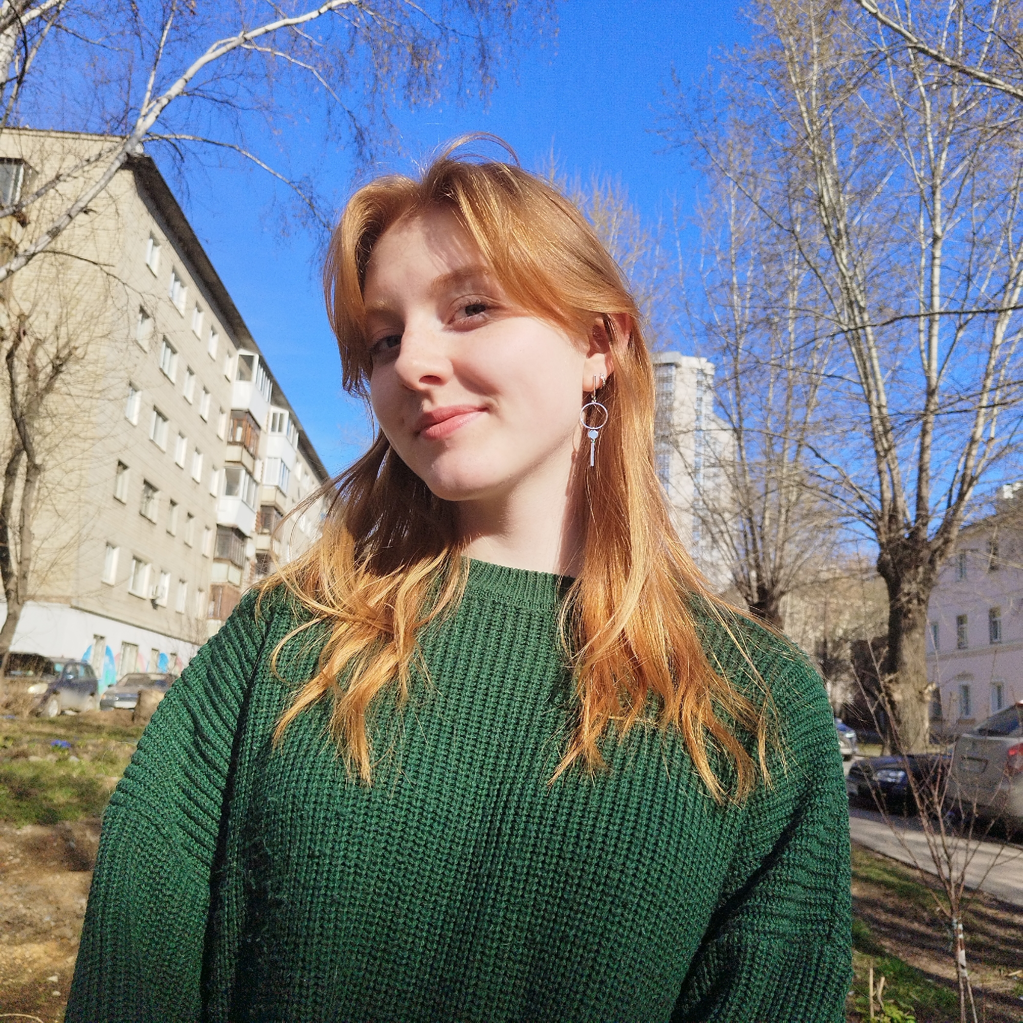 A young woman with reddish-blonde hair and light skin smiles gently while looking directly at the camera. She is wearing a dark green, ribbed knitted sweater and a silver-colored dangle earring with a small light-colored bead on her right ear. Her hair is parted in the middle and falls over her shoulders.
The setting is outdoors on a bright, sunny day, indicated by a clear blue sky and strong light on the subject. The background features an urban or suburban residential area. To the left, a light-colored, multi-story apartment building with many windows and balconies is visible. Numerous bare trees, possibly birches due to their pale bark, are scattered throughout the background, their leafless branches reaching towards the sky. On the right side, more bare trees and a distant building can be seen, along with what appears to be a street with parked cars further back. The ground is covered with sparse grass or dry earth. The absence of leaves on the trees suggests the photo was taken in late autumn, winter, or early spring. No visible text is present in the image.