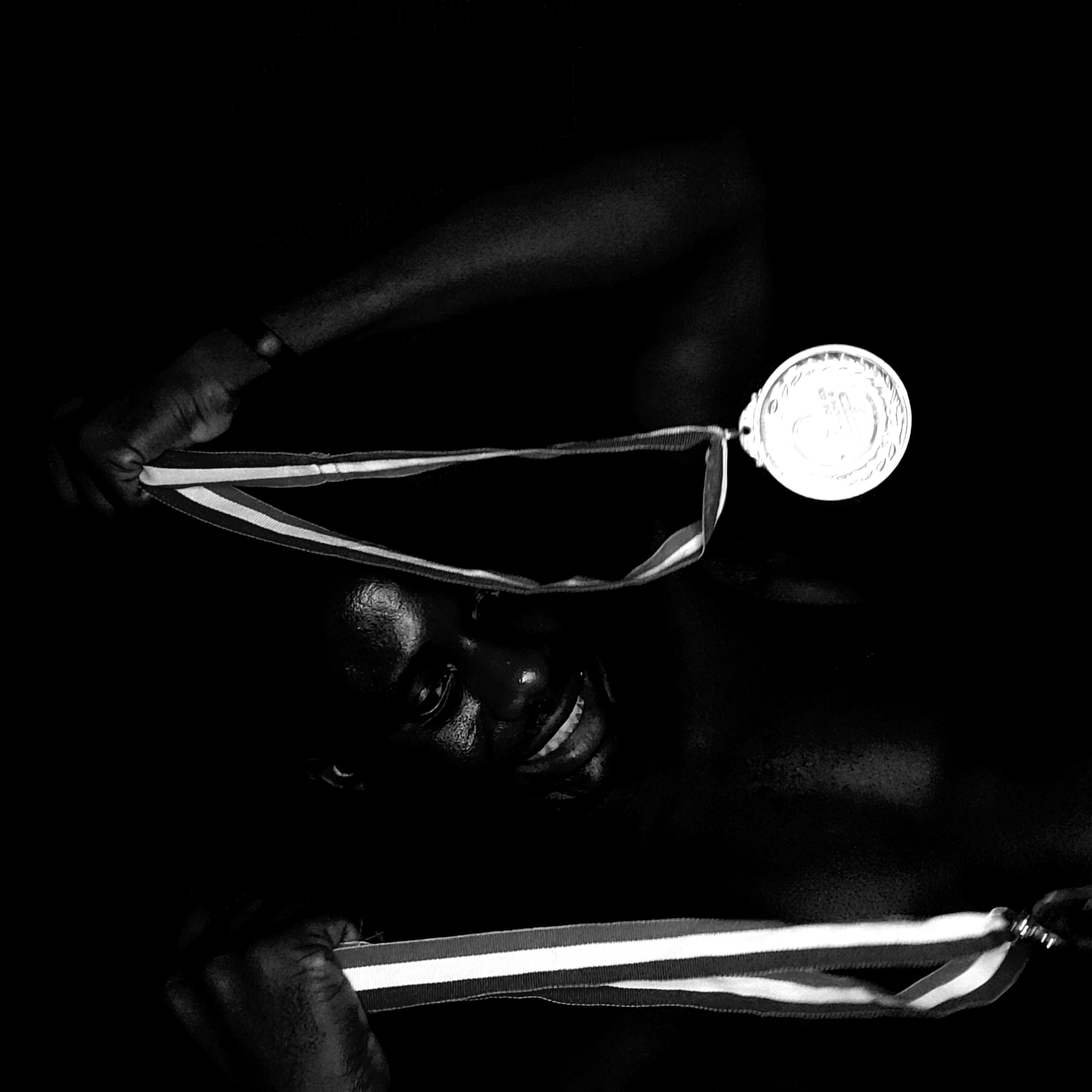 This striking black and white, high-contrast image captures a moment of joy and accomplishment. The central subject is a person, likely male, with dark skin, whose face emerges from a completely dark background, illuminated by a strong light source. A broad, joyful smile is prominent on their face, with visible teeth and glistening skin, suggesting either sweat from exertion or intentional lighting effects.
The individual is holding a medal with a striped ribbon (featuring red, white, and dark stripes) with both hands. The hands are positioned on either side of the medal's ribbon, holding it up. The circular medal itself is brightly lit and reflective, appearing to be metallic (possibly silver or gold). It displays intricate embossed details but no clearly legible text.
The setting is minimalist, with the deep black background serving to isolate and emphasize the subject and the medal. This creates a dramatic and intimate atmosphere, focusing solely on the individual's expression of pride and achievement. The overall impression is one of triumph and happiness, against the implied context of Narayi, Nigeria.