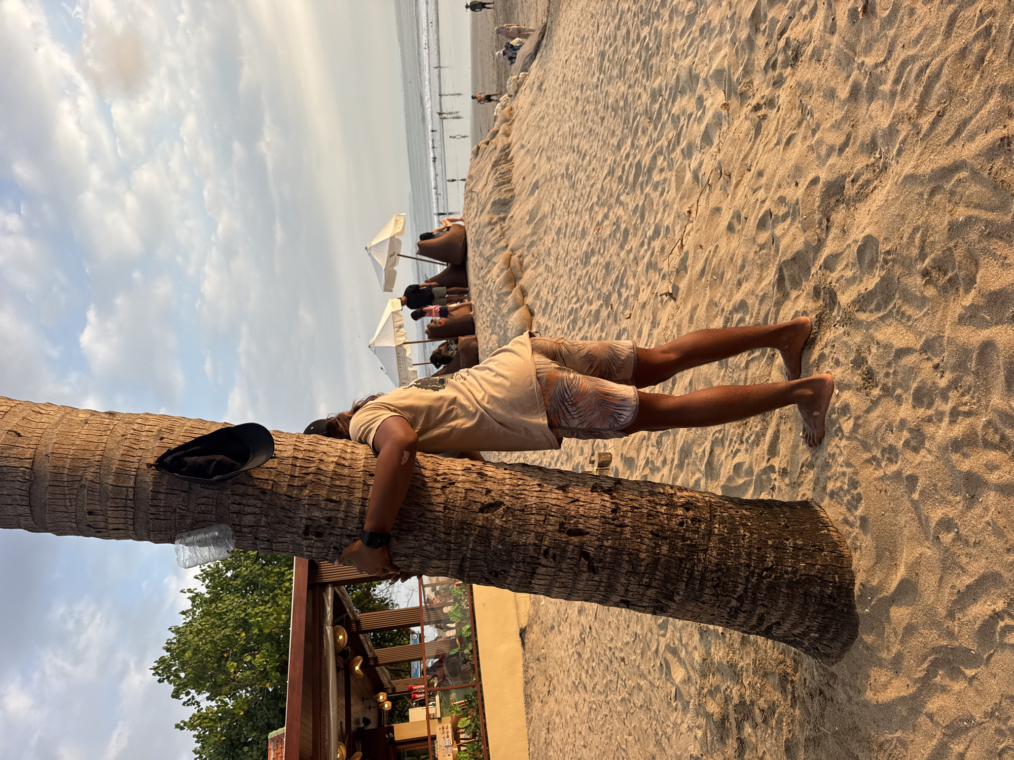 A person is hugging a large palm tree on a sandy beach in Kuta, Indonesia, at sunset. The person is wearing a beige t-shirt and patterned shorts, and a black baseball cap is hanging from the tree. To the left of the tree is a beachfront building with tables and chairs, and in the background, the ocean can be seen with people walking along the shore and in the water. White umbrellas and beanbag chairs are set up on a sand dune. The sky is cloudy with hints of orange and yellow from the setting sun.