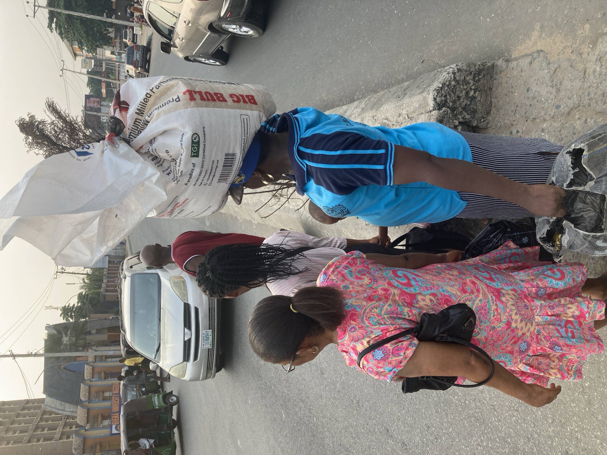 The image depicts a street scene, likely in Aba, Nigeria, given the vehicle license plate marked "Abia" and the architecture of the buildings in the background. A person is carrying a large sack labeled "BIG BULL Premium Milled Parboiled Rice" on their head. They are wearing a blue shirt with dark blue stripes on the sleeves. Next to them, several people appear to be crouched or kneeling near the curb, with a parked silver Toyota Sienna minivan beside them. One person is wearing a pink and blue floral dress, while another is wearing a red shirt. A tricycle (Keke) can be seen in the background on the left side of the image. Overhead power lines are also visible, and the overall lighting suggests it is daytime.